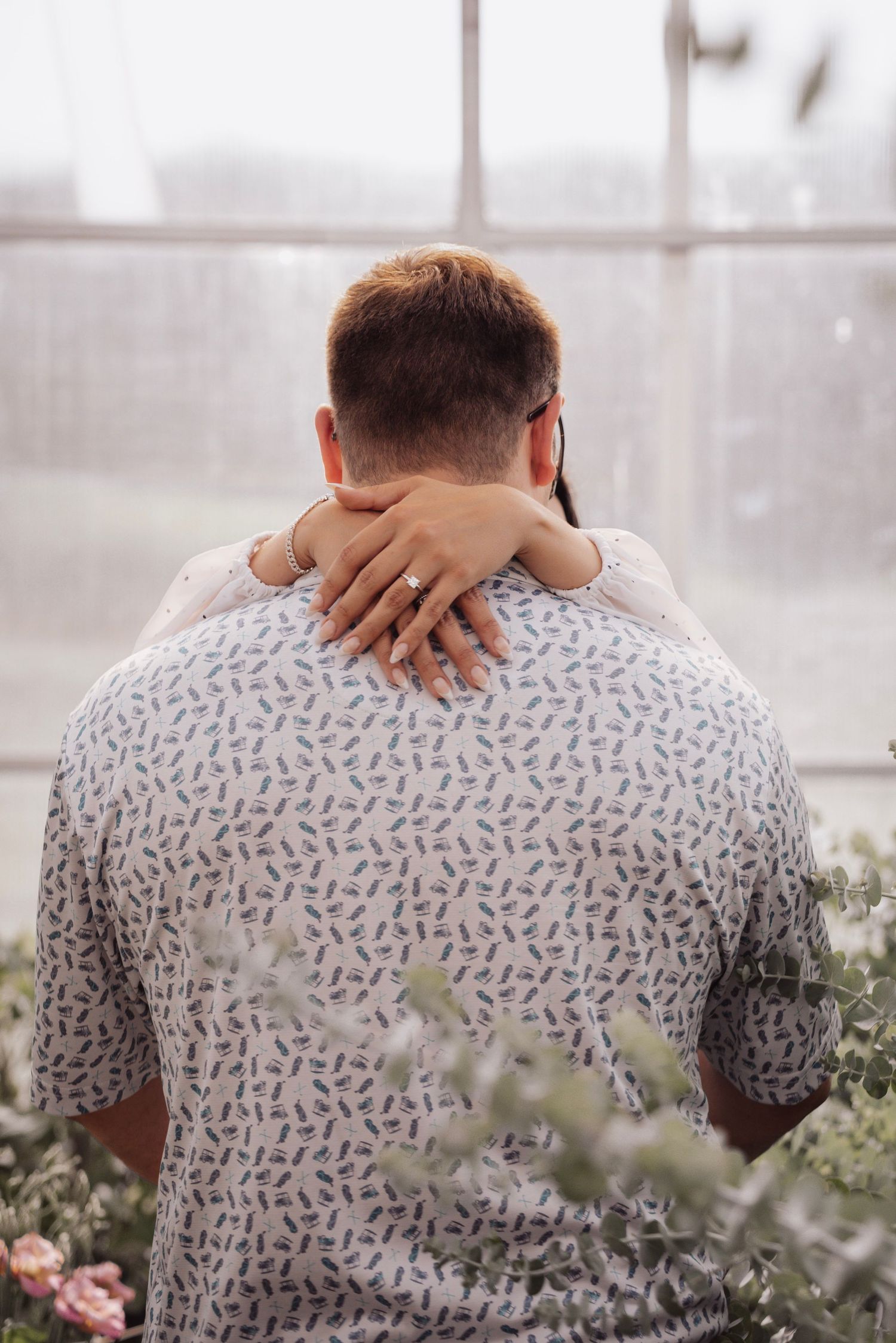 Back view of a person wearing a patterned shirt sitting on a bench near plants in a bright indoor space.