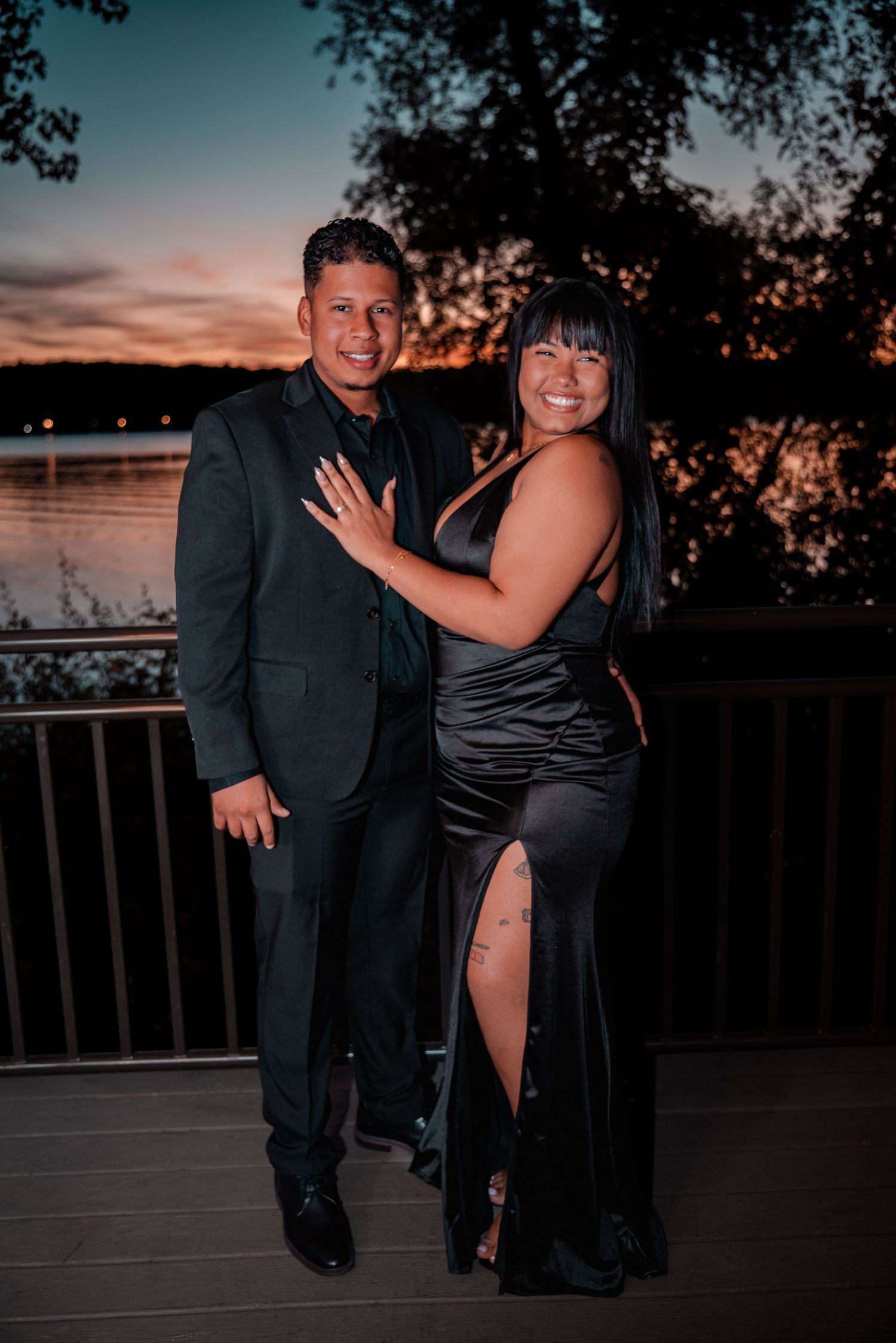 Couple in formal attire dancing and posing together at sunset on a waterfront deck.