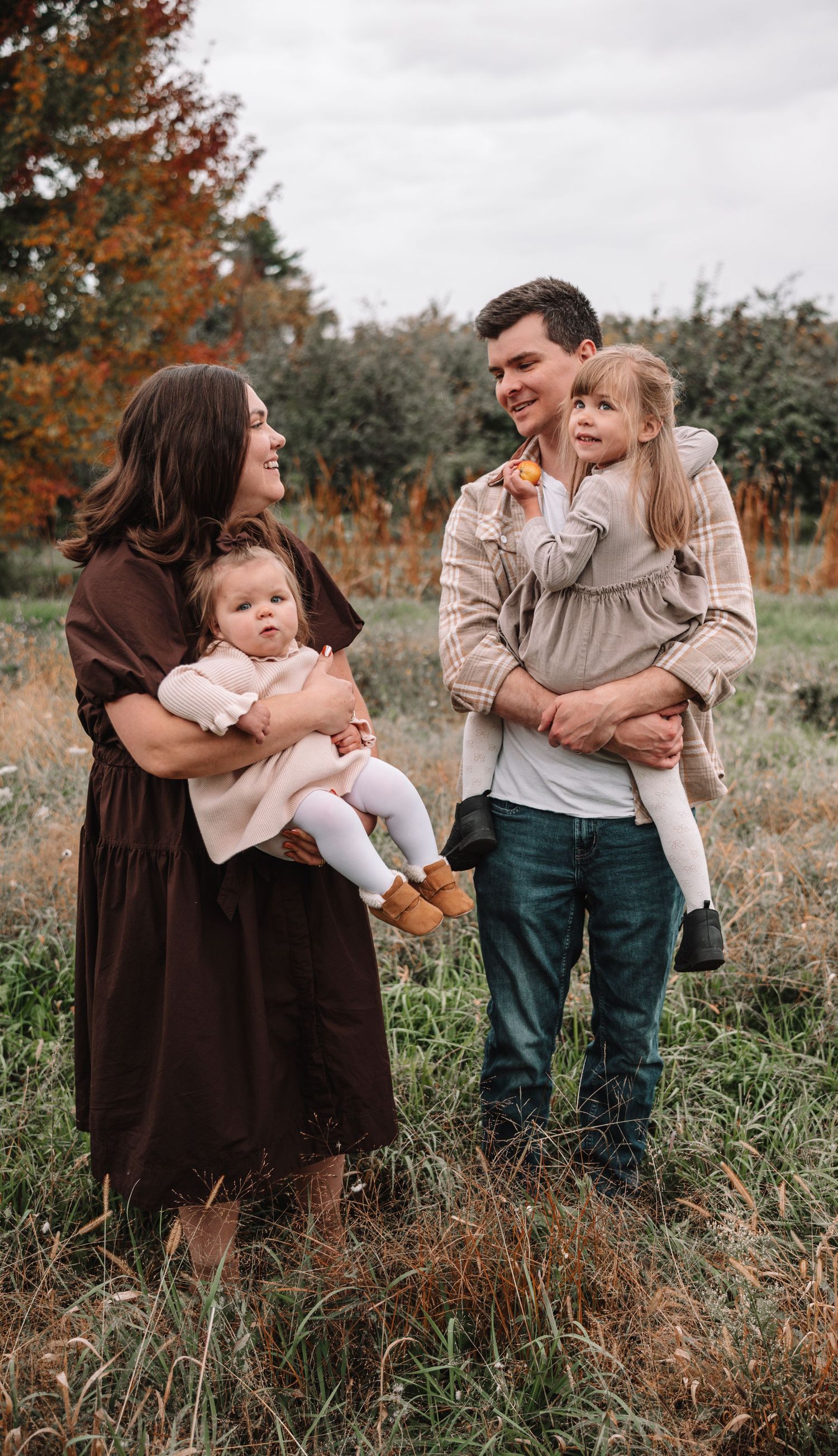A family shares tender moments together in a field during autumn, wearing cozy neutral-toned clothing.