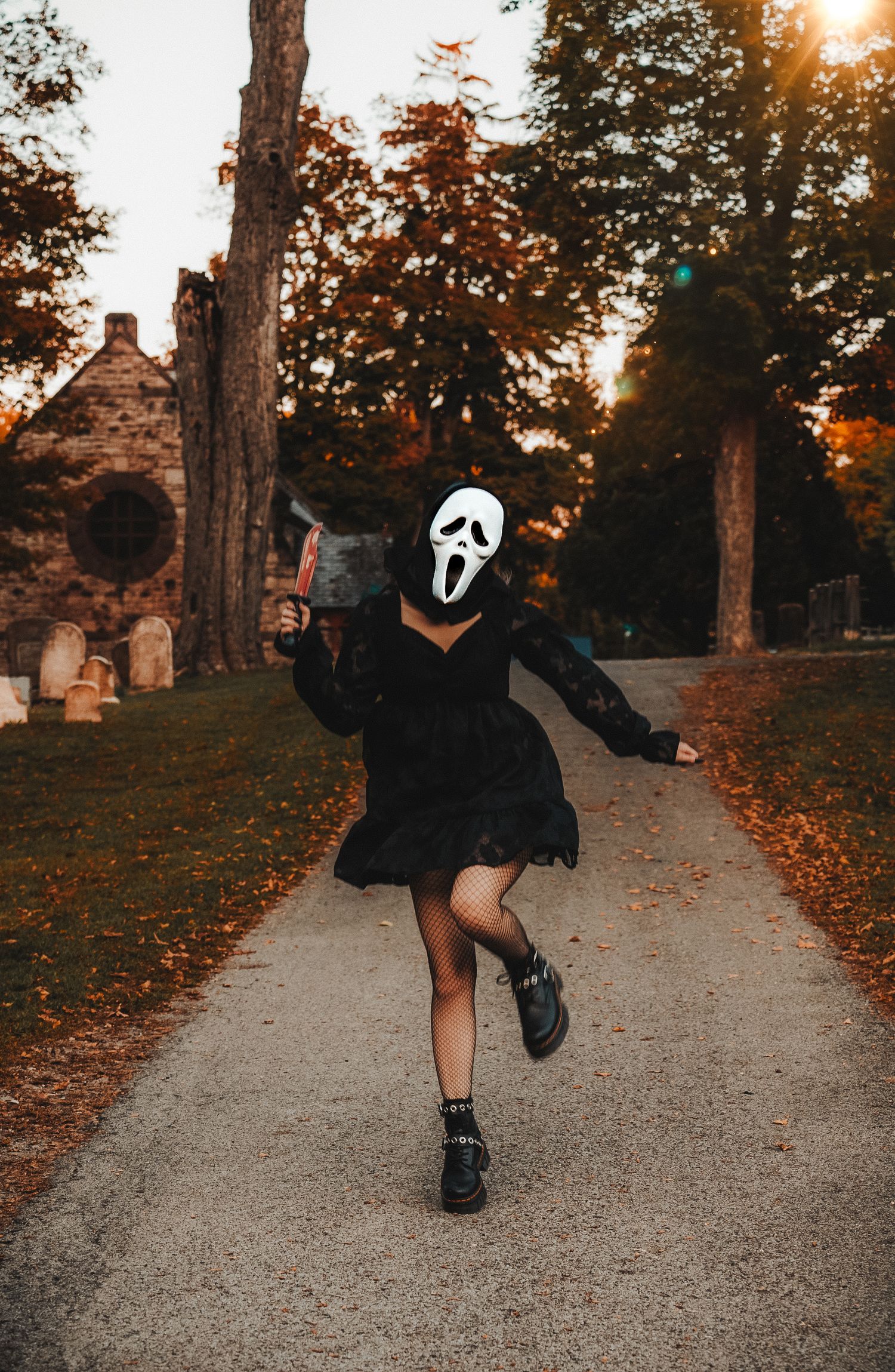 A figure in a dark outfit and ghost mask performs dance moves on a cemetery path at sunset.