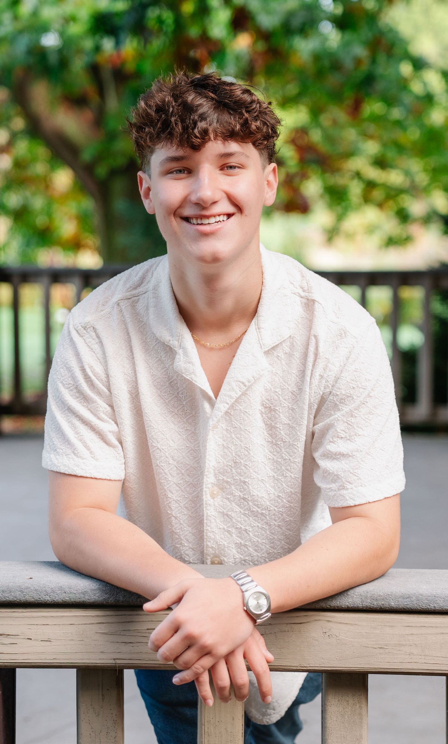 Outdoor portrait of a young person in a white collared shirt sitting at a wooden railing with greenery in background.