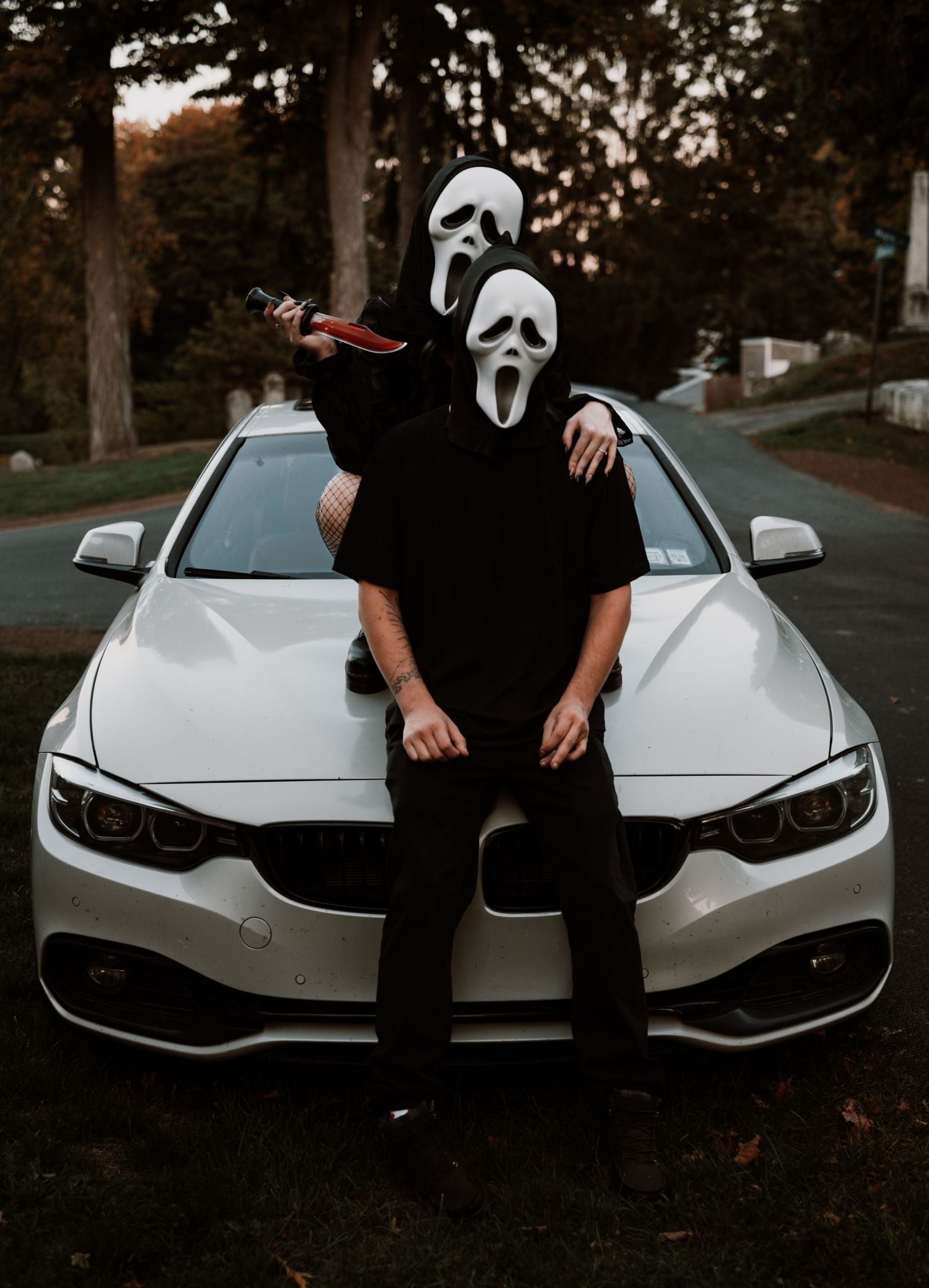 Figure in Ghostface Scream mask poses in front of white BMW car at dusk wearing all black clothing.