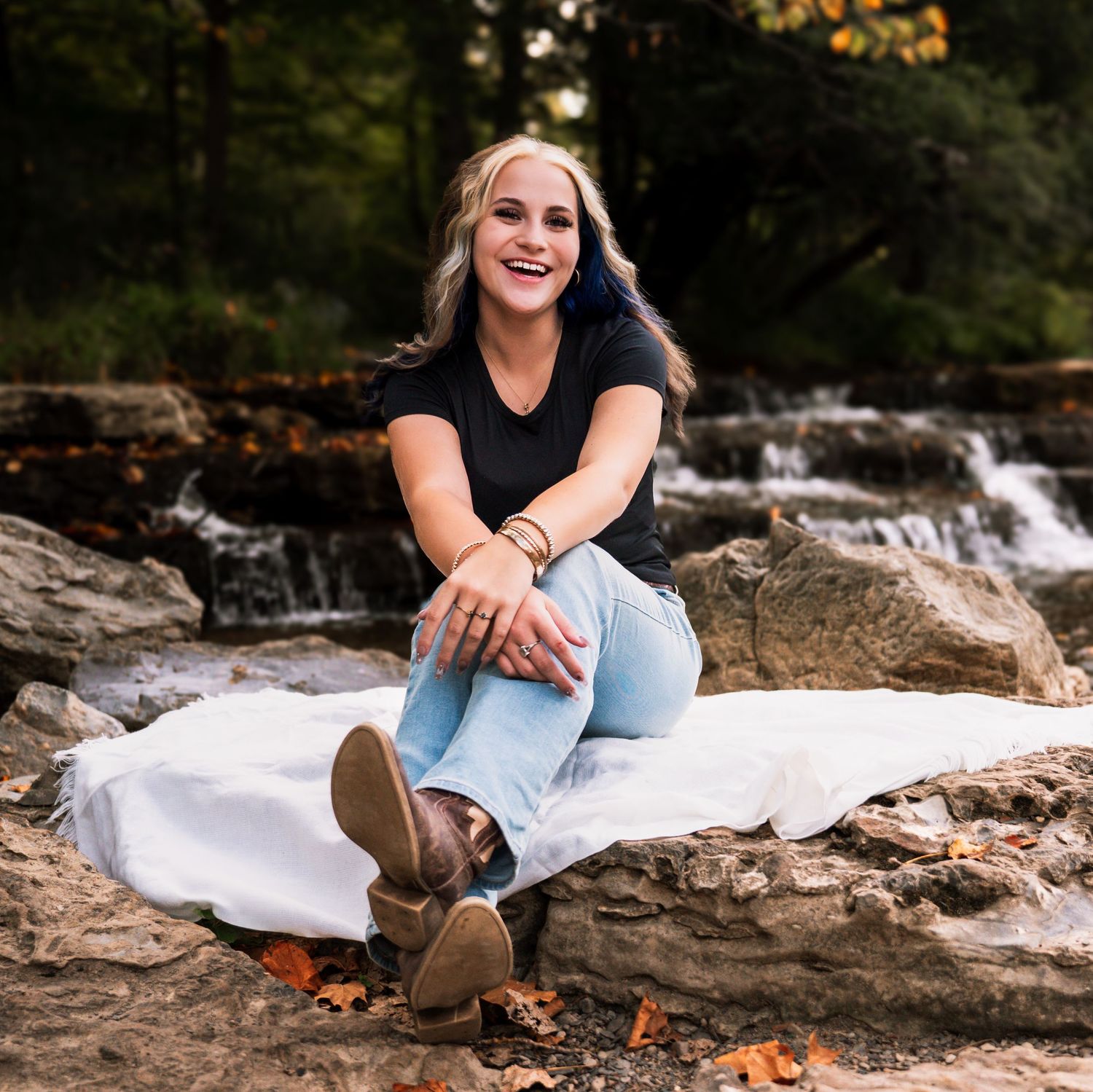 A person sits casually on a white blanket by a cascading waterfall on rocky terrain wearing boots and casual attire.