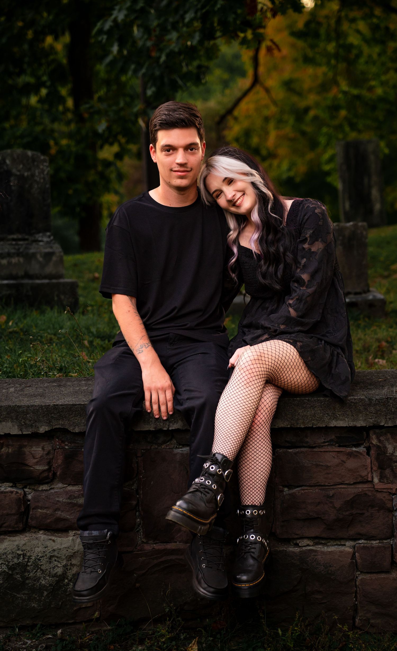 A couple in black outfits sits intimately on stone steps in a moody outdoor setting during golden hour.