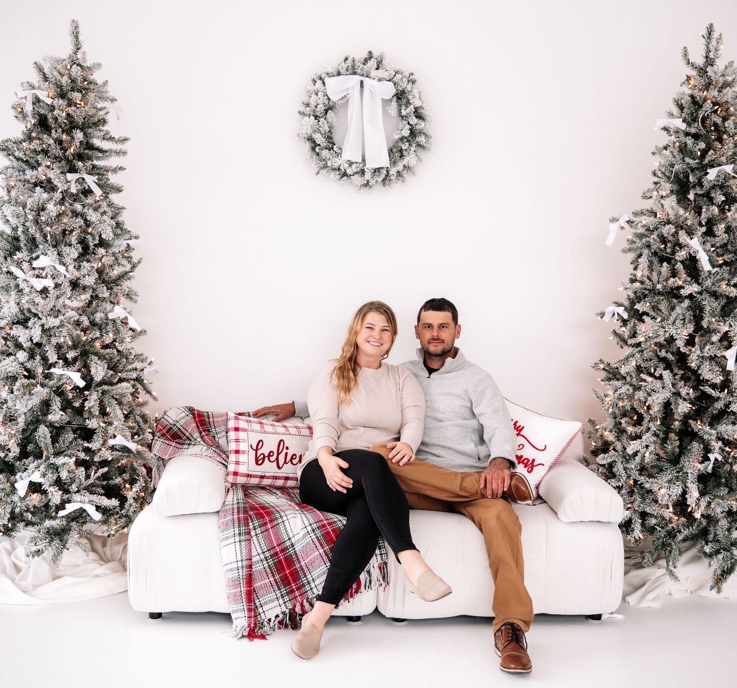 A cozy holiday scene with two people sitting on a white couch between snow-covered Christmas trees and a wreath on the wall.