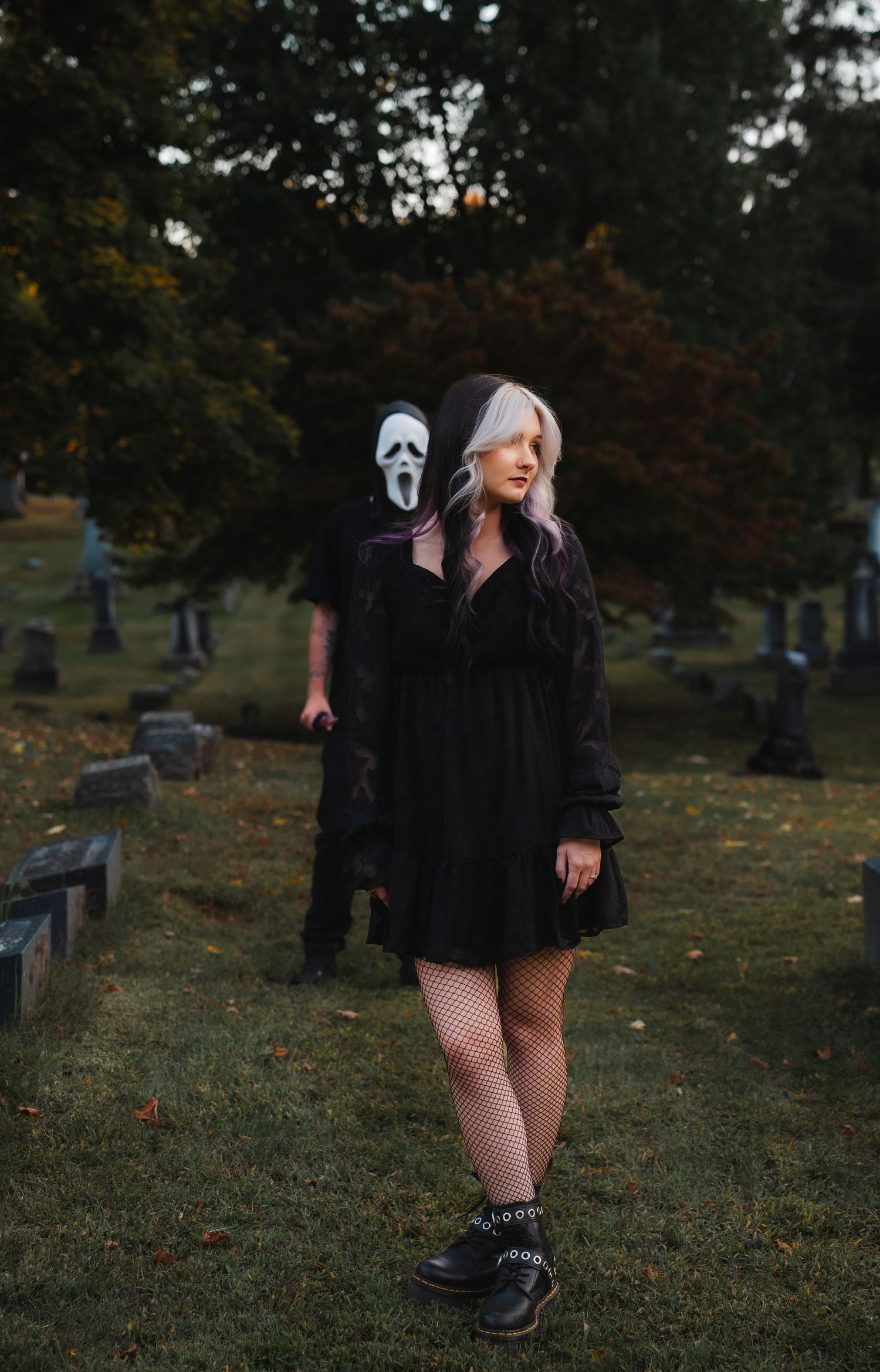 A figure in black clothing and fishnet stockings poses in a graveyard during autumn.