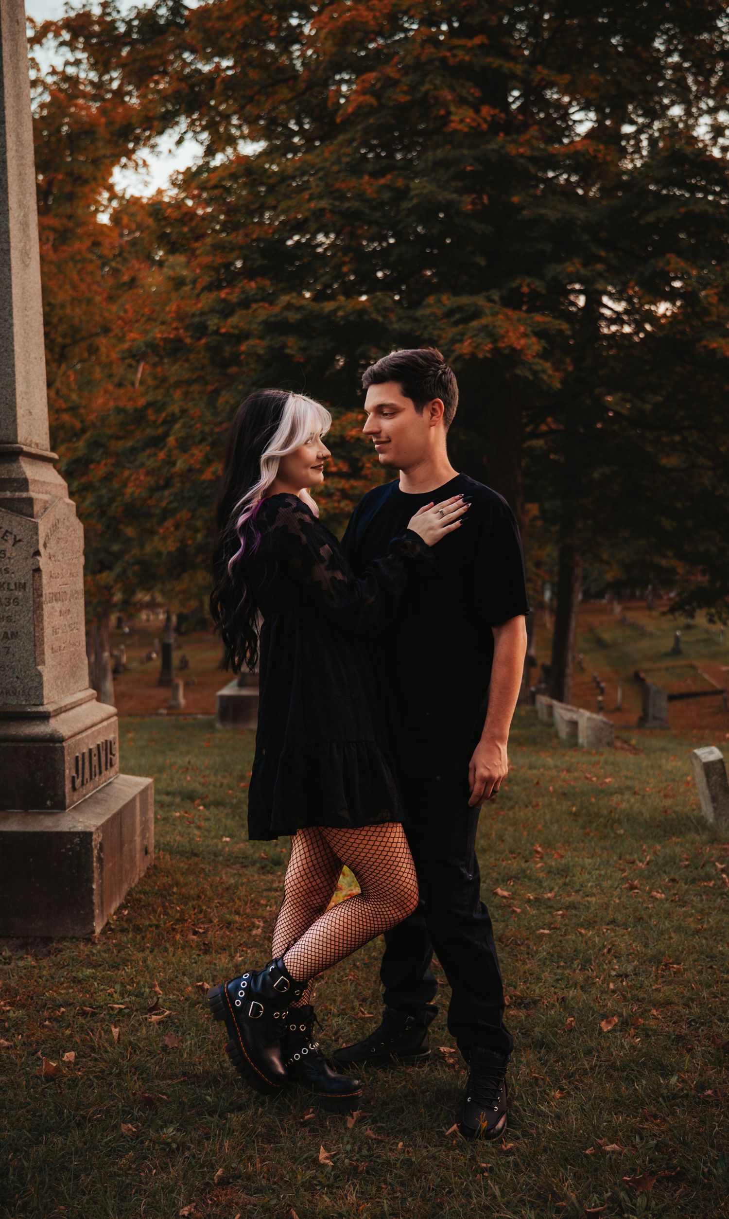 A romantic couple in dark clothing embraces near a stone monument in an autumn cemetery with fallen leaves.