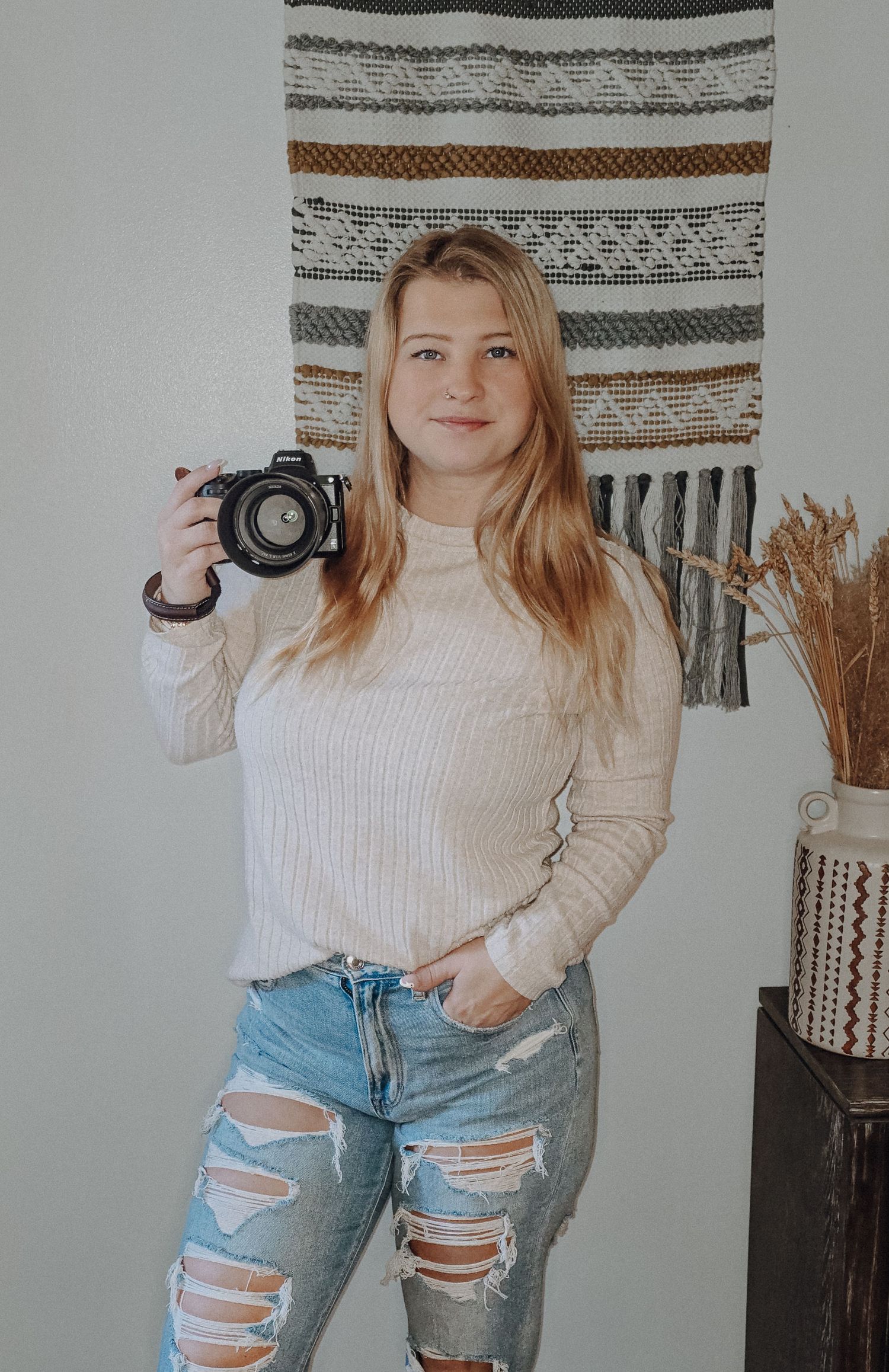 Person in cream sweater and distressed jeans holds camera against white wall with striped tapestry decor.