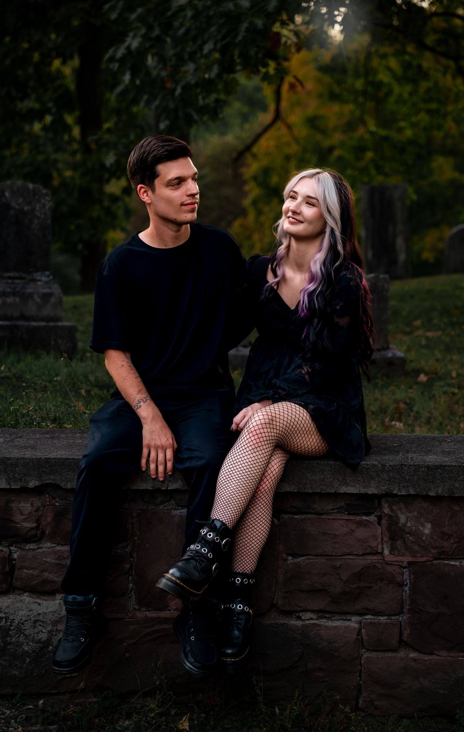A couple sits intimately on a stone wall at dusk surrounded by lush greenery and autumn foliage.