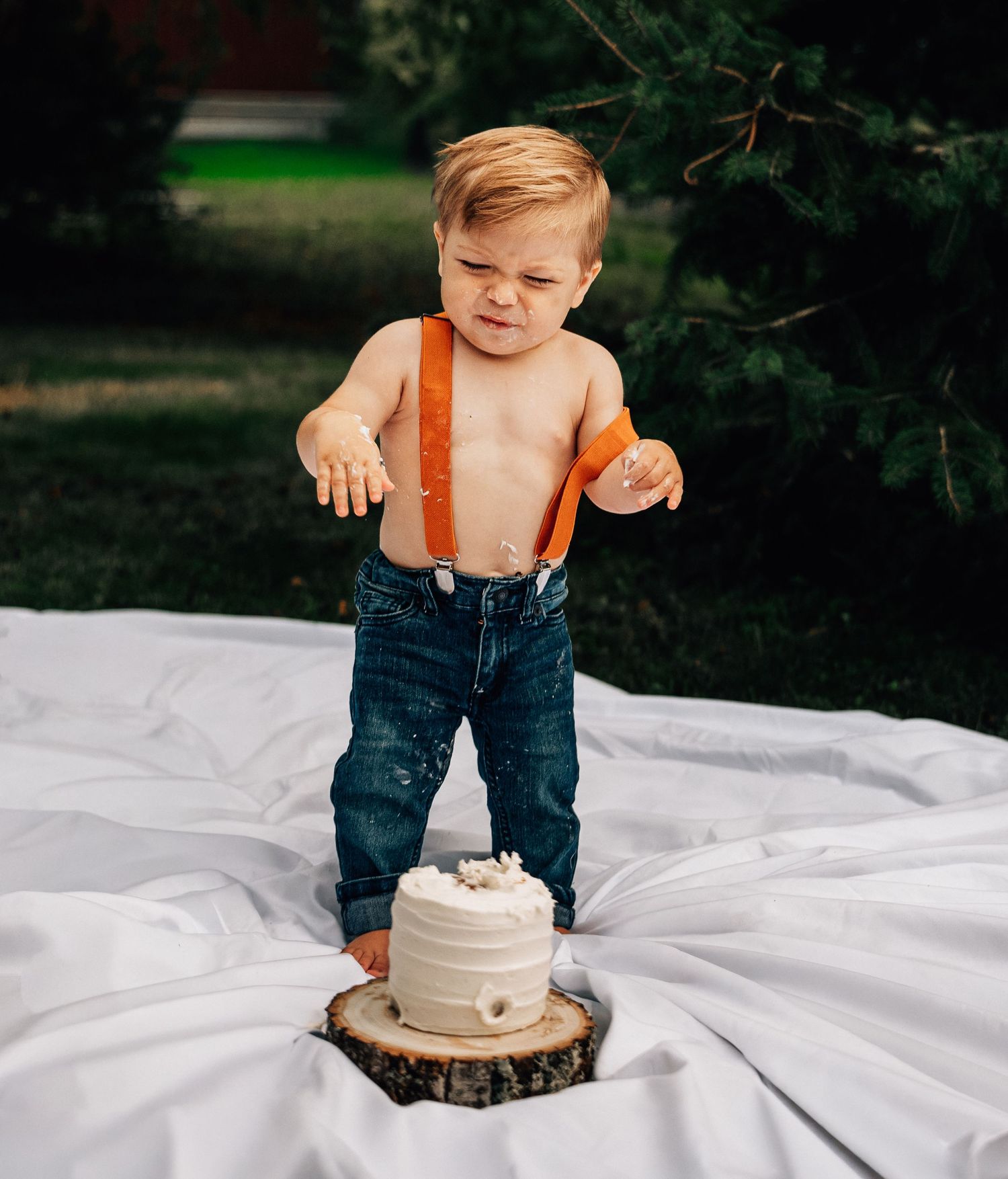 Small child in jeans and orange suspenders stands on white blanket with rustic cake display during outdoor photoshoot.