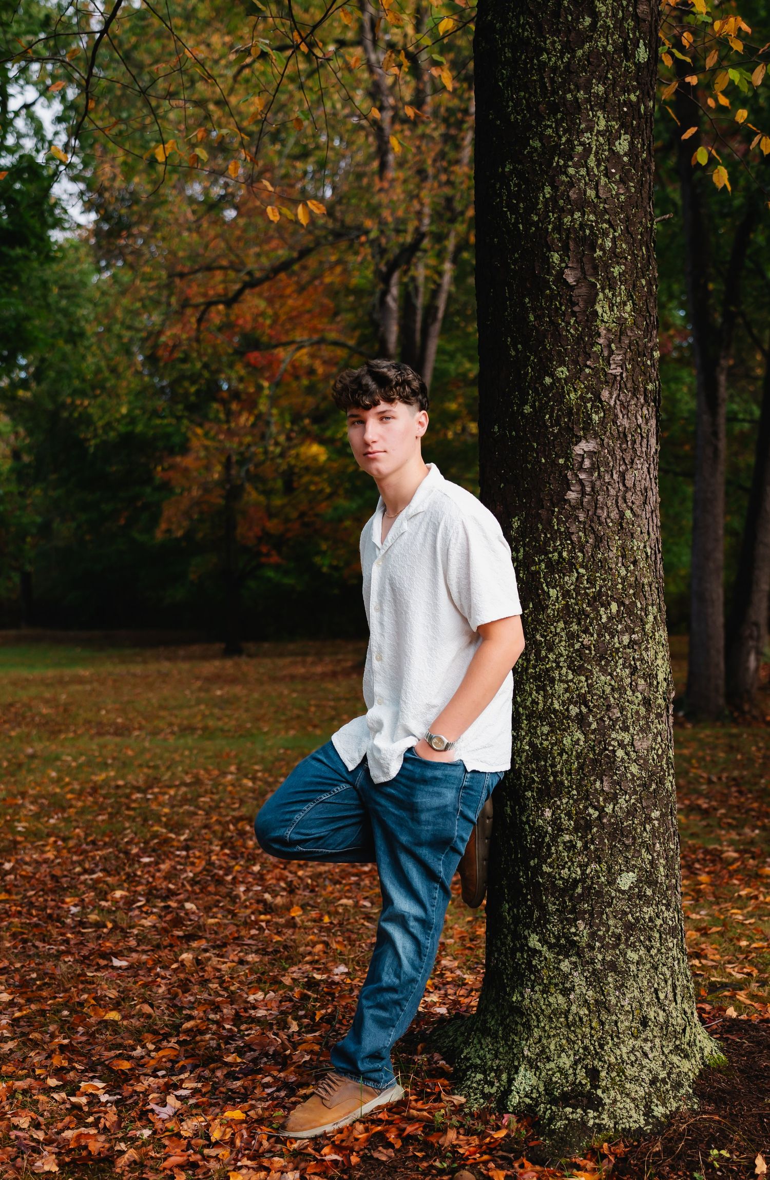 A person in a white shirt and jeans leans against a tree trunk surrounded by autumn leaves in a park setting.