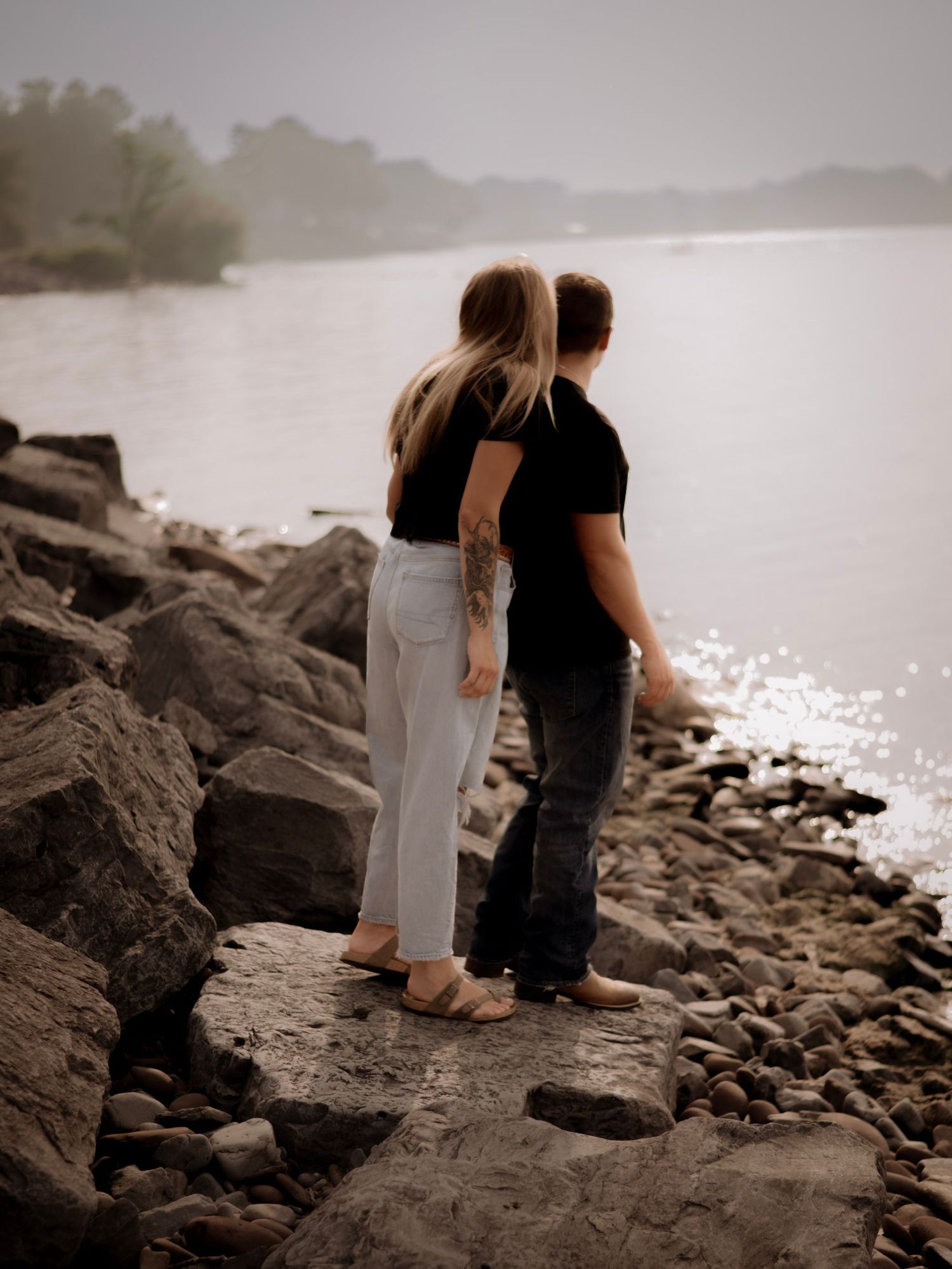 A couple sits together on rocky coastal shoreline at sunset creating a romantic silhouette against the water.
