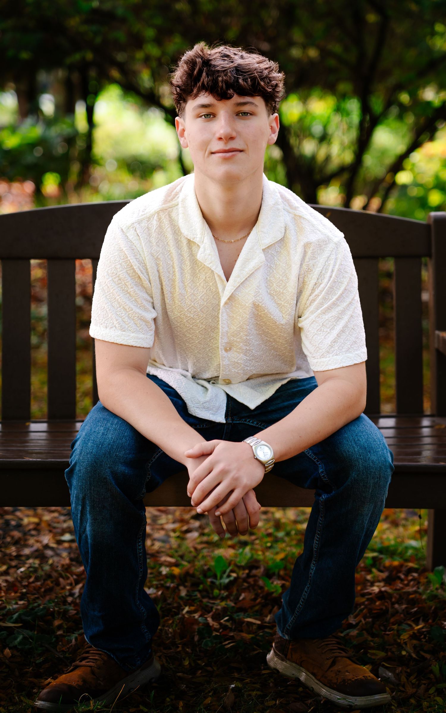 Portrait of someone in a white button-down shirt and jeans sitting on a wooden bench in an outdoor garden setting.