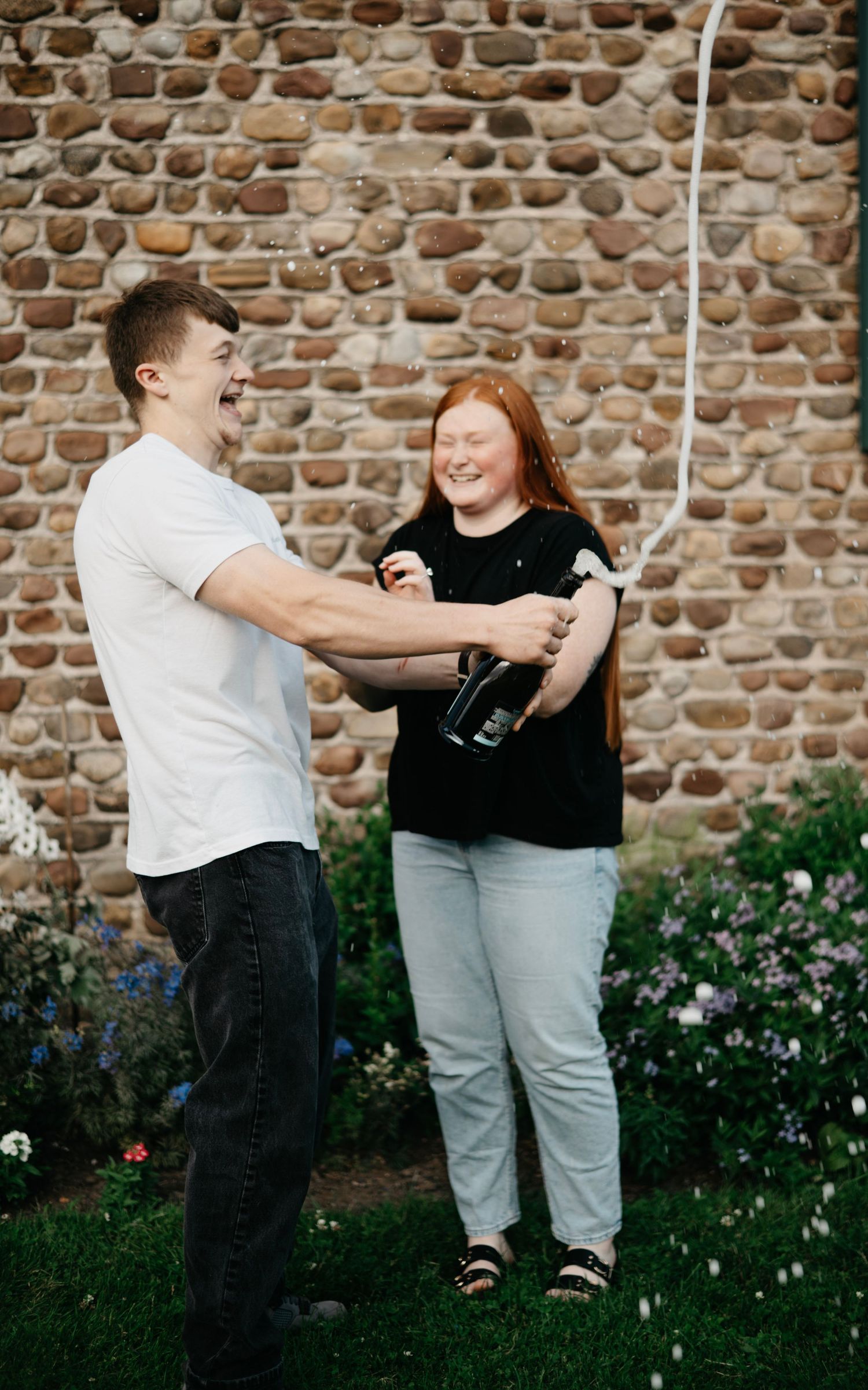 Couple enjoying playful moments in front of a rustic brick wall with flowering garden.