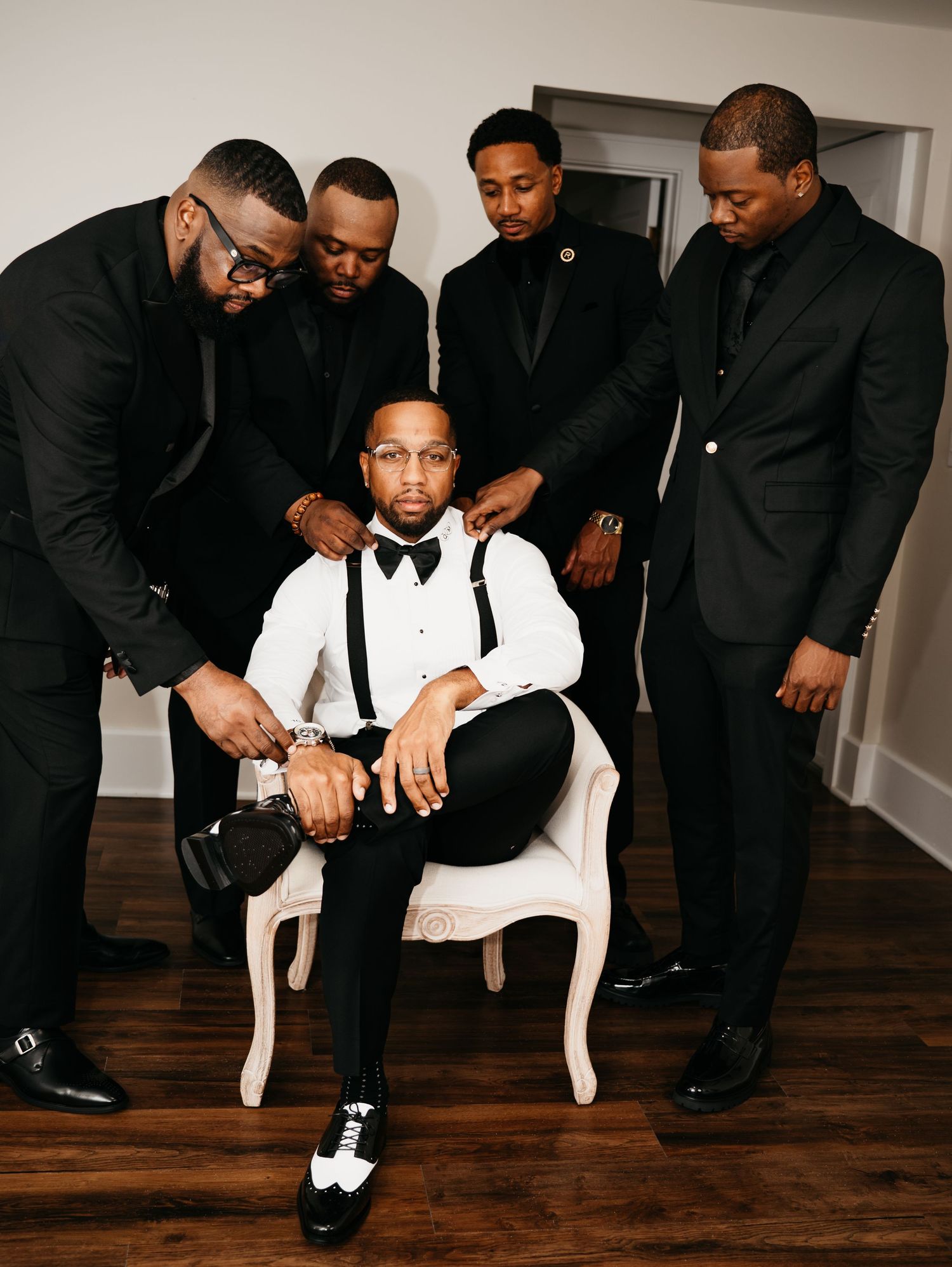 A group of men in formal black suits gather around another in white and black attire seated in an elegant chair.