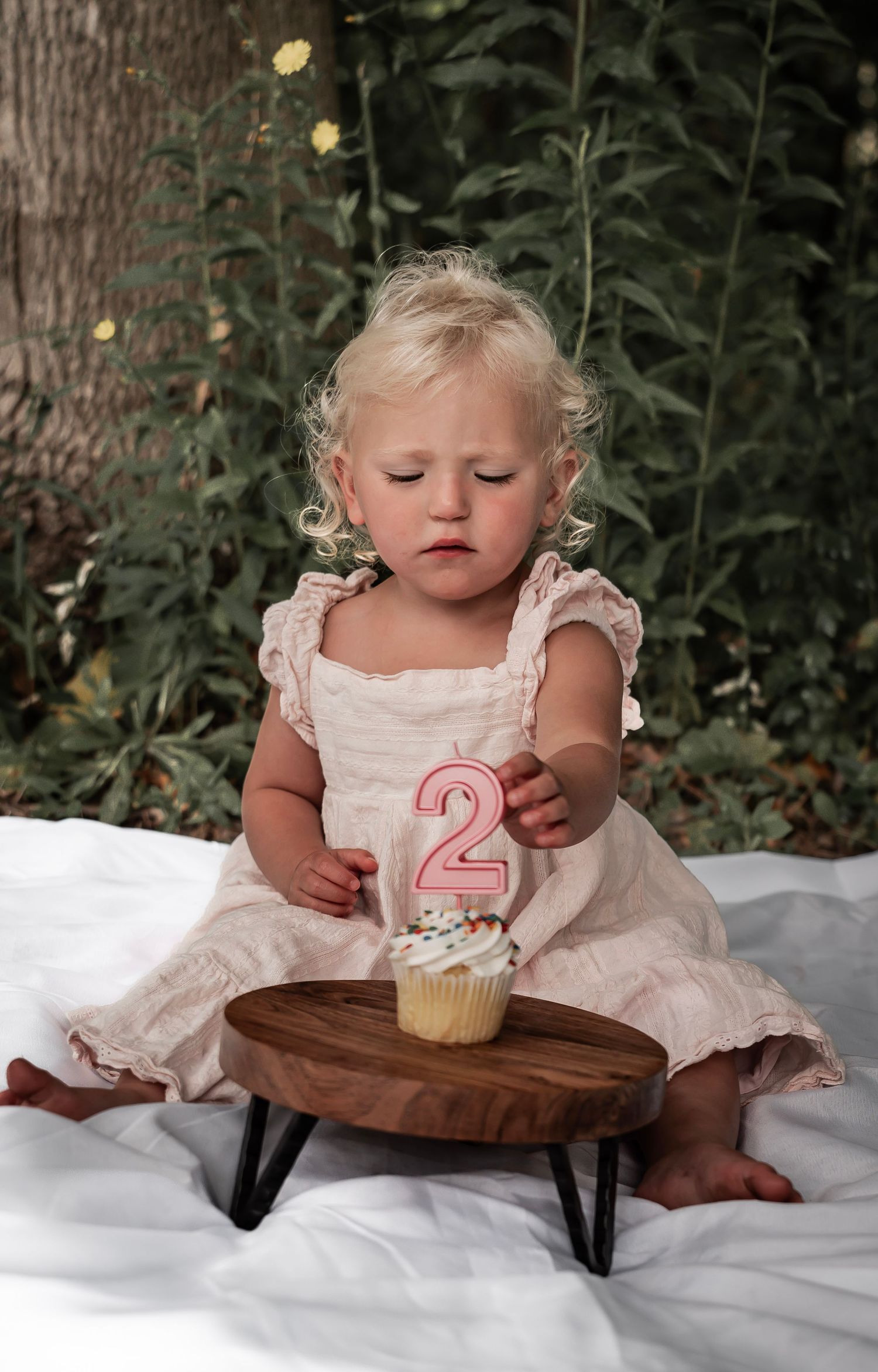 A toddler in a white dress sits on a blanket with a cupcake decorated with a number 2 candle.