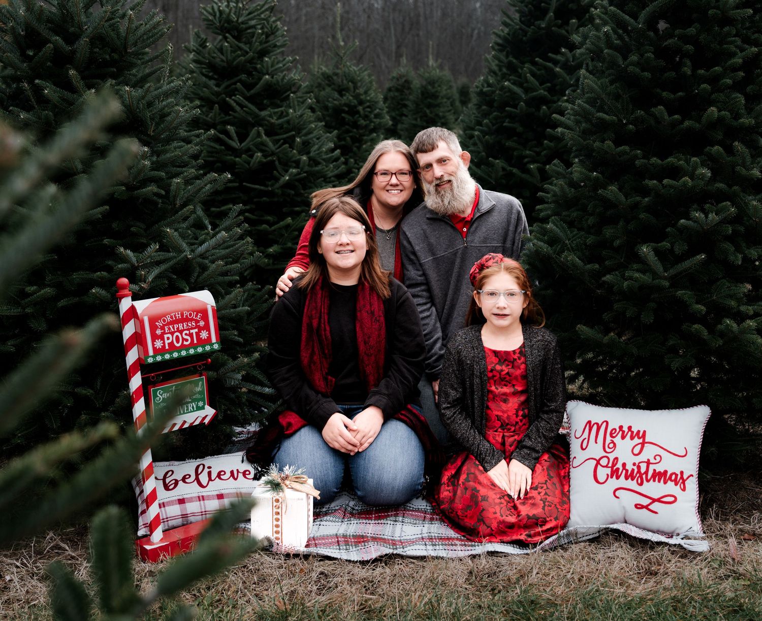 People sit among evergreen trees during a winter photo session with red and white holiday decor and blankets.