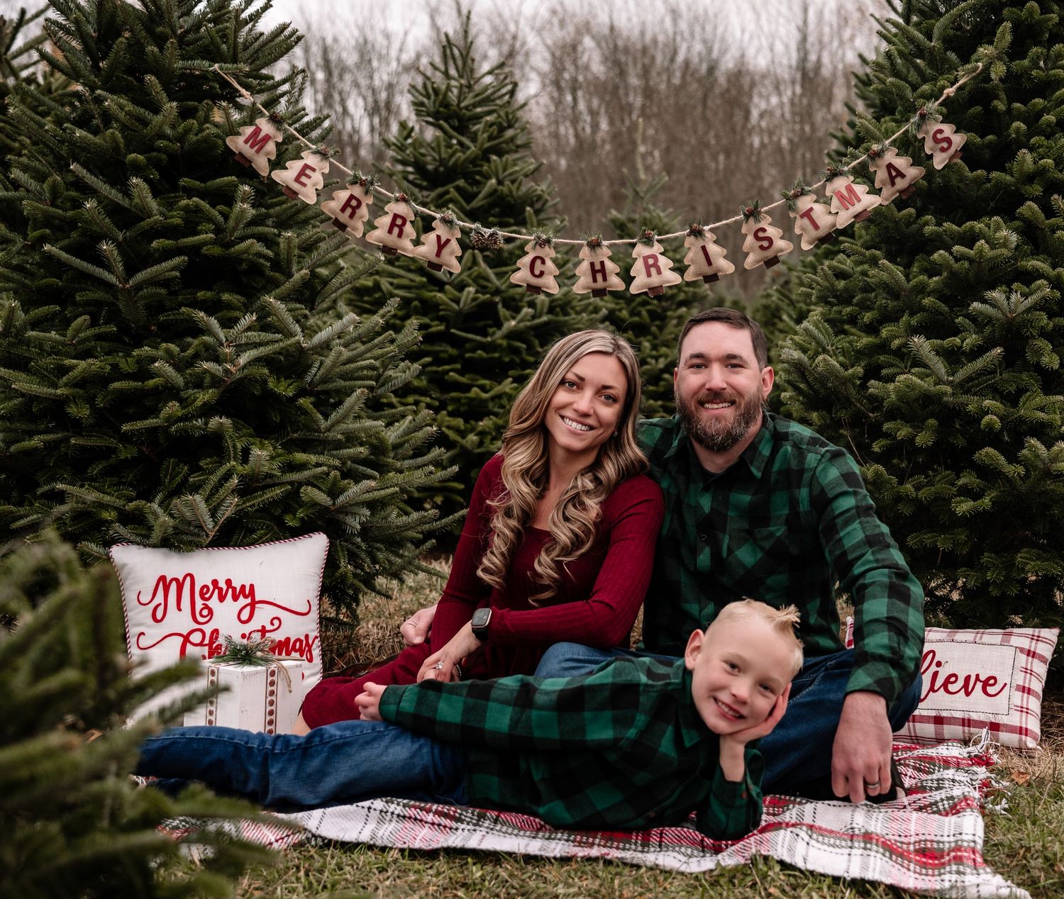 Family poses together on blanket at Christmas tree farm with festive holiday decorations and signs.