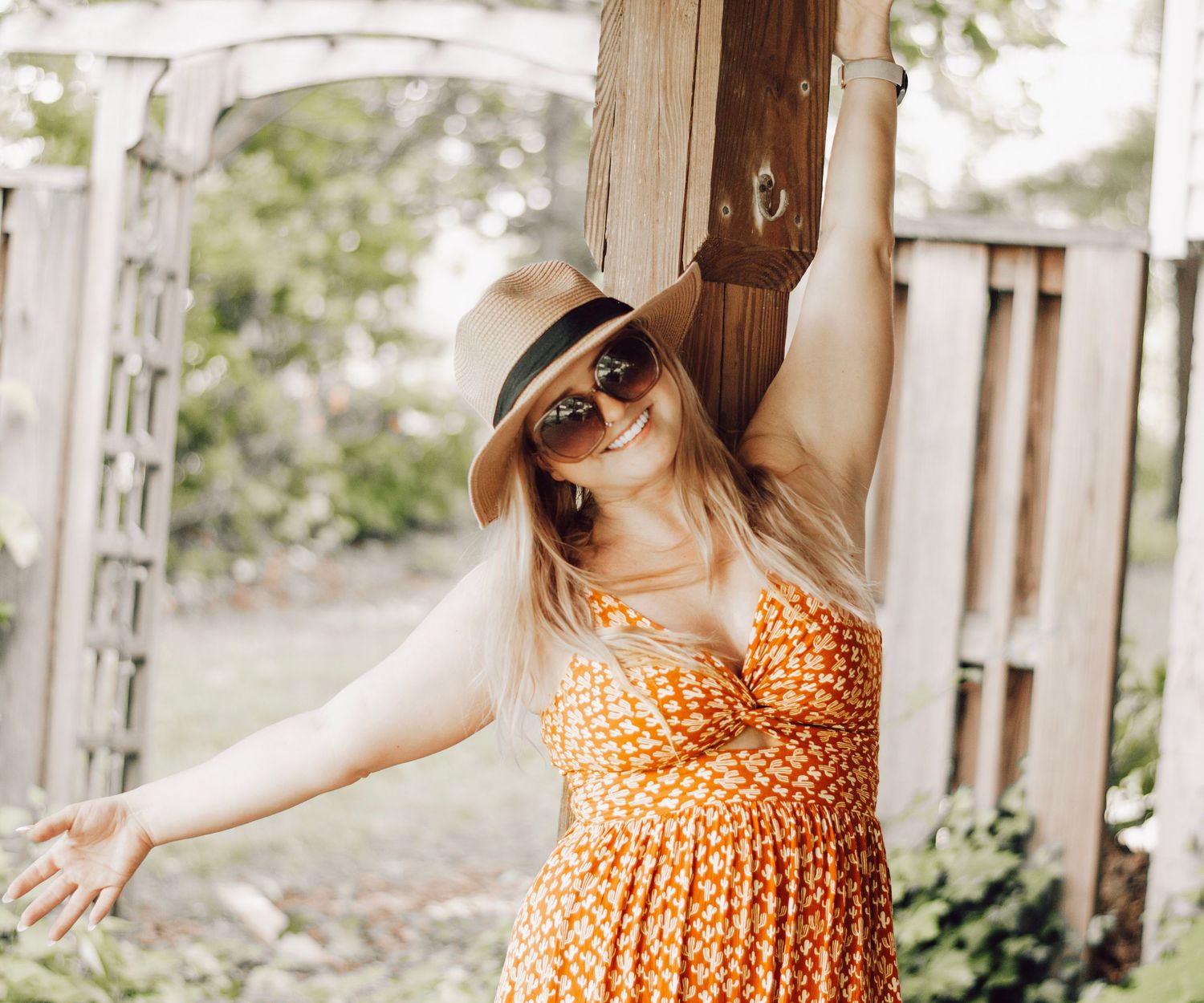 Woman in orange dress and hat poses playfully on garden pergola.
