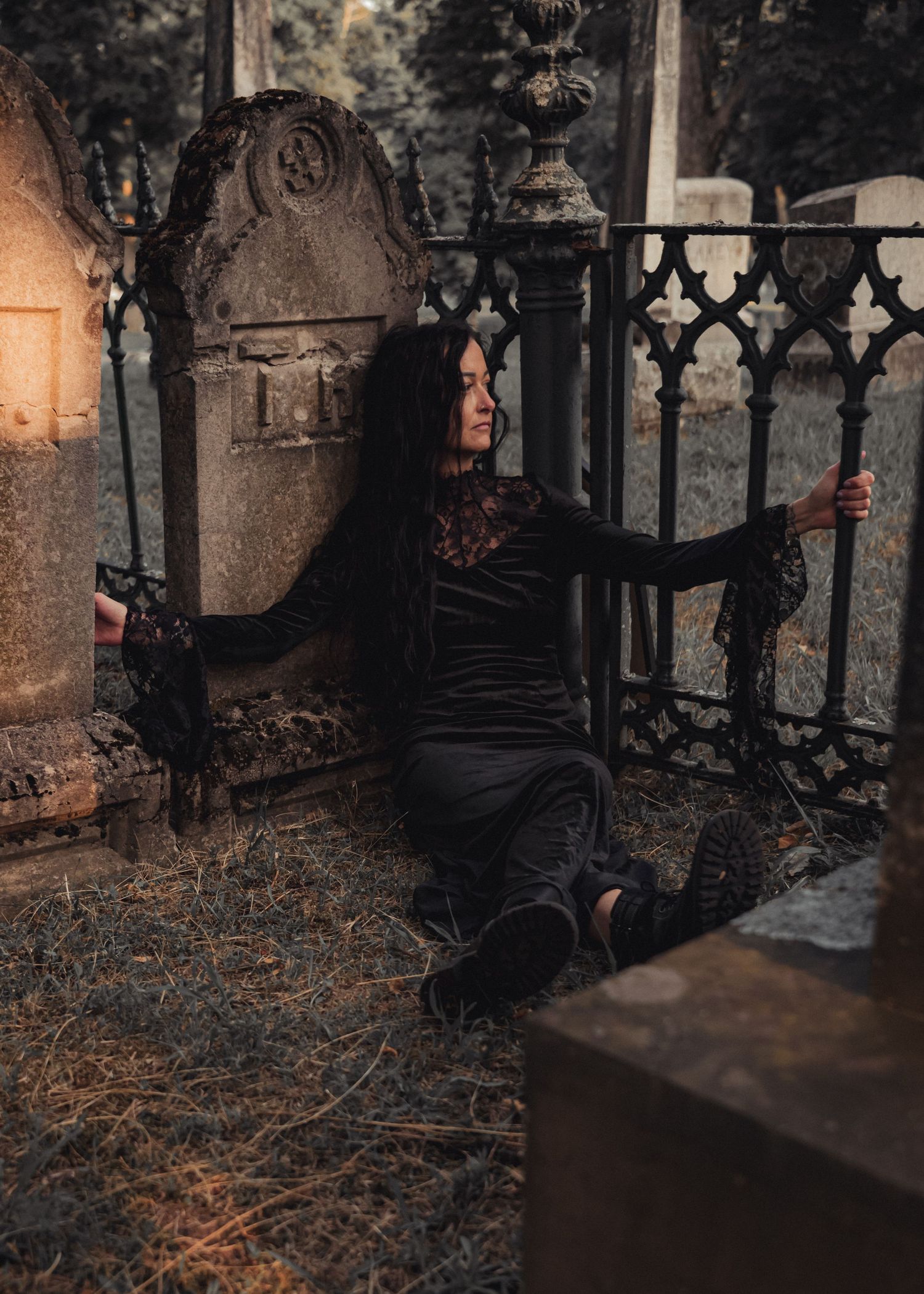 A gothic scene shows a figure in dark clothing sitting between ornate iron cemetery gates at dusk.