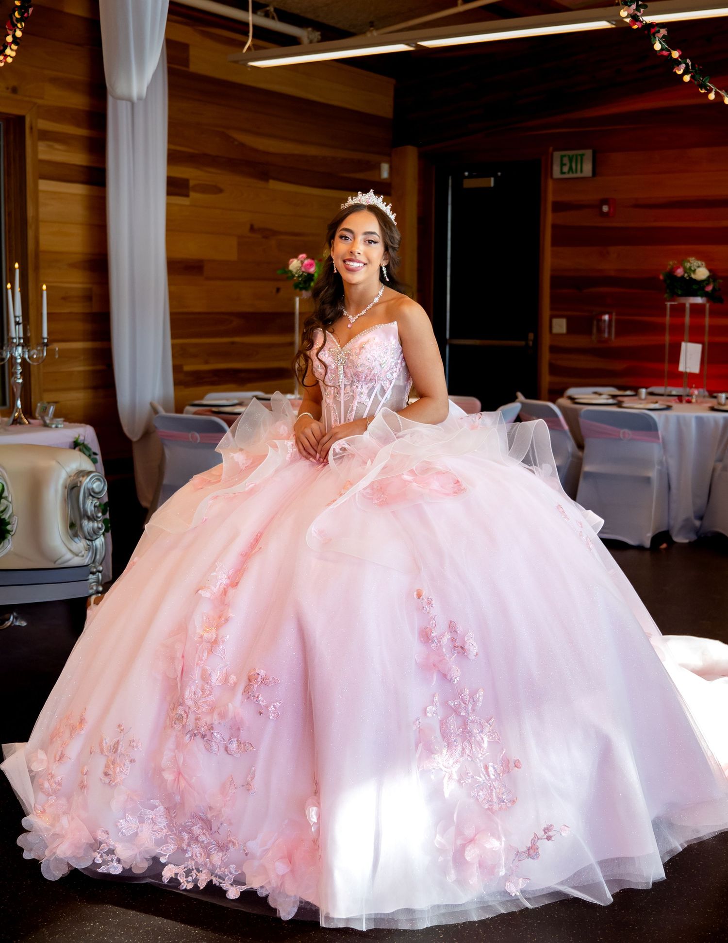 A young woman in a pink and white quinceañera dress poses gracefully in an elegant reception hall setting.