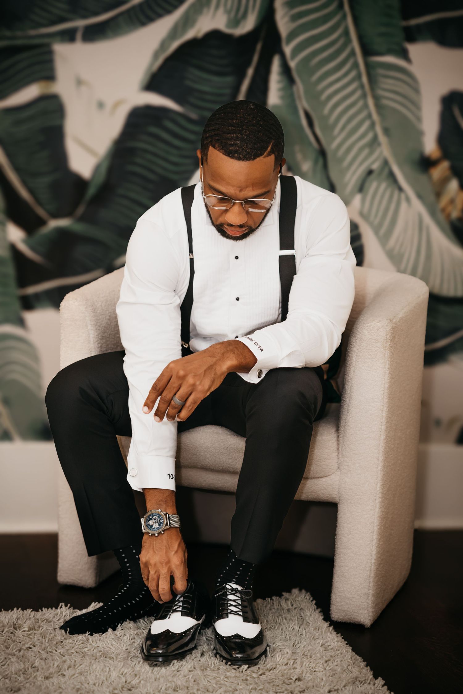 A formally dressed person in white shirt and suspenders sits in a gray armchair tying two-tone dress shoes.