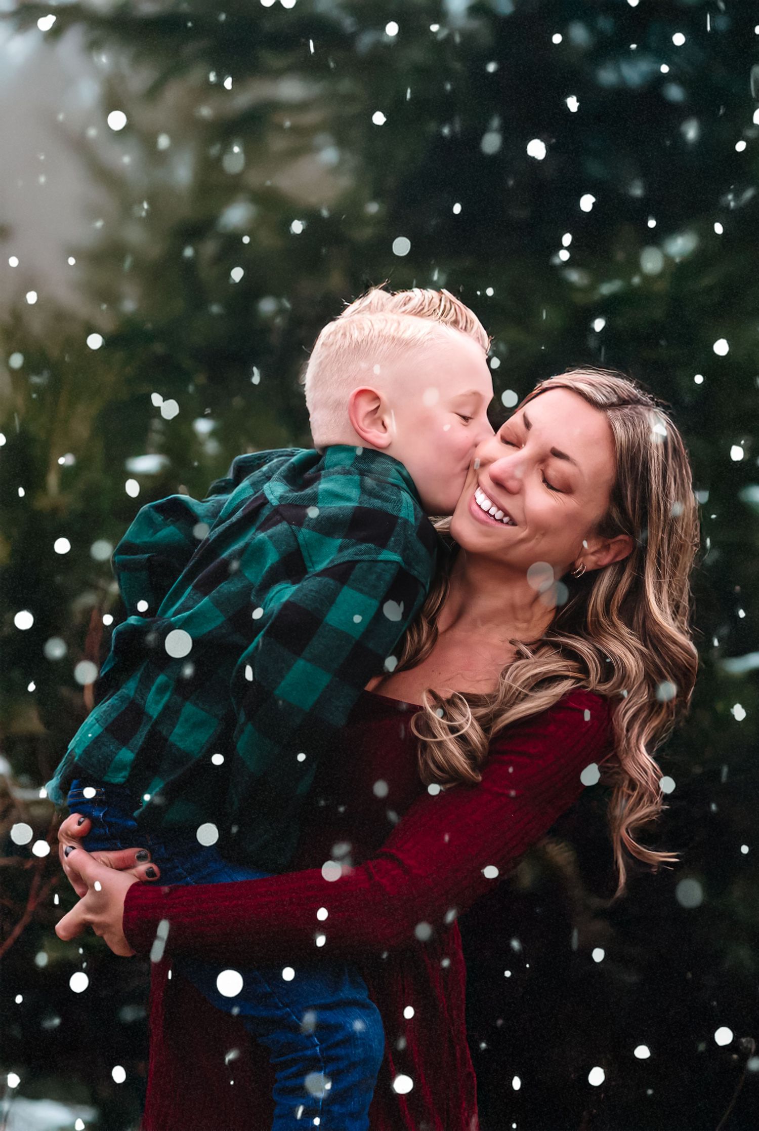 A couple shares a romantic moment in falling snow while wearing a plaid shirt and burgundy sweater.
