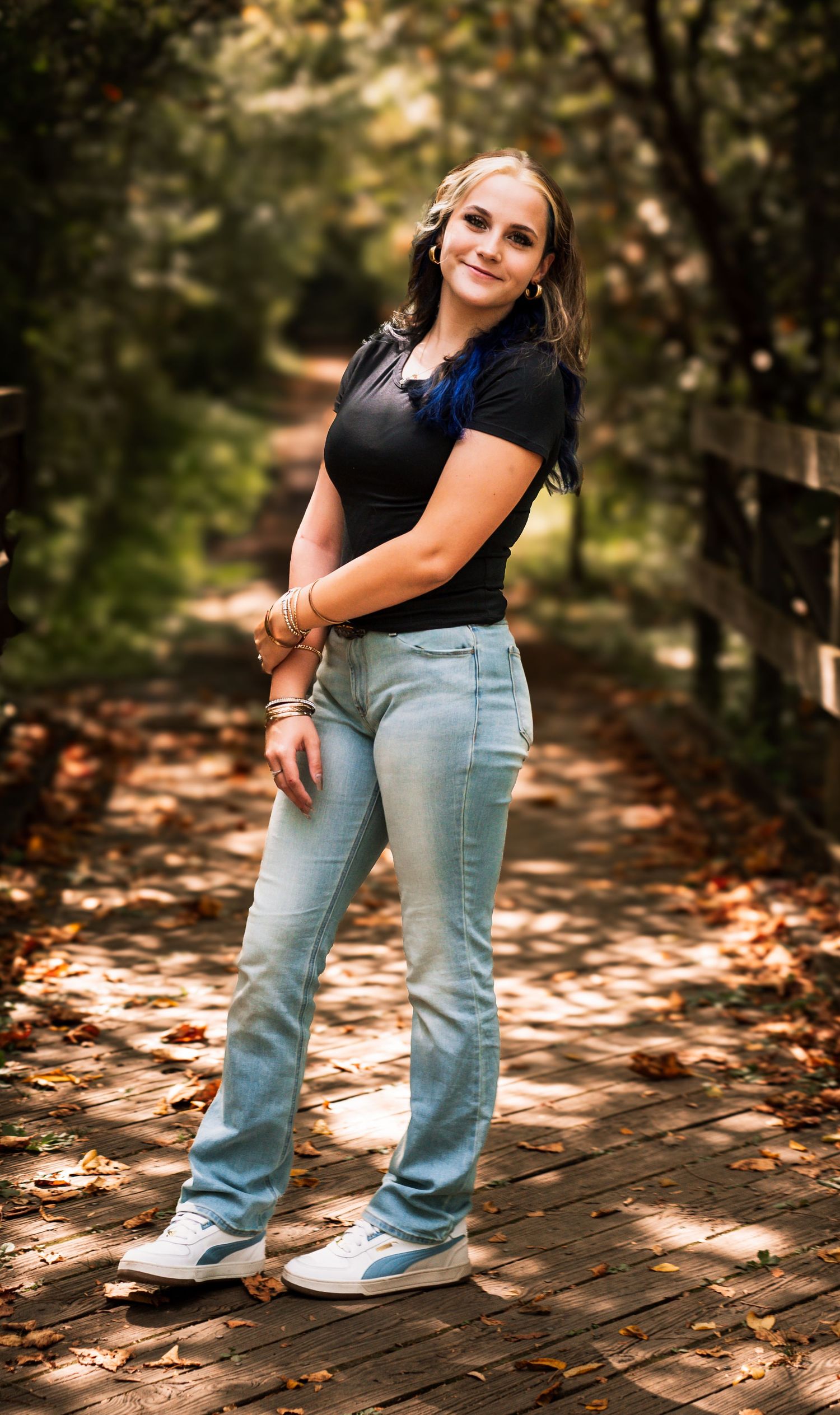 Person wearing light blue jeans and black top poses playfully on a leaf-covered brick path in dappled sunlight.
