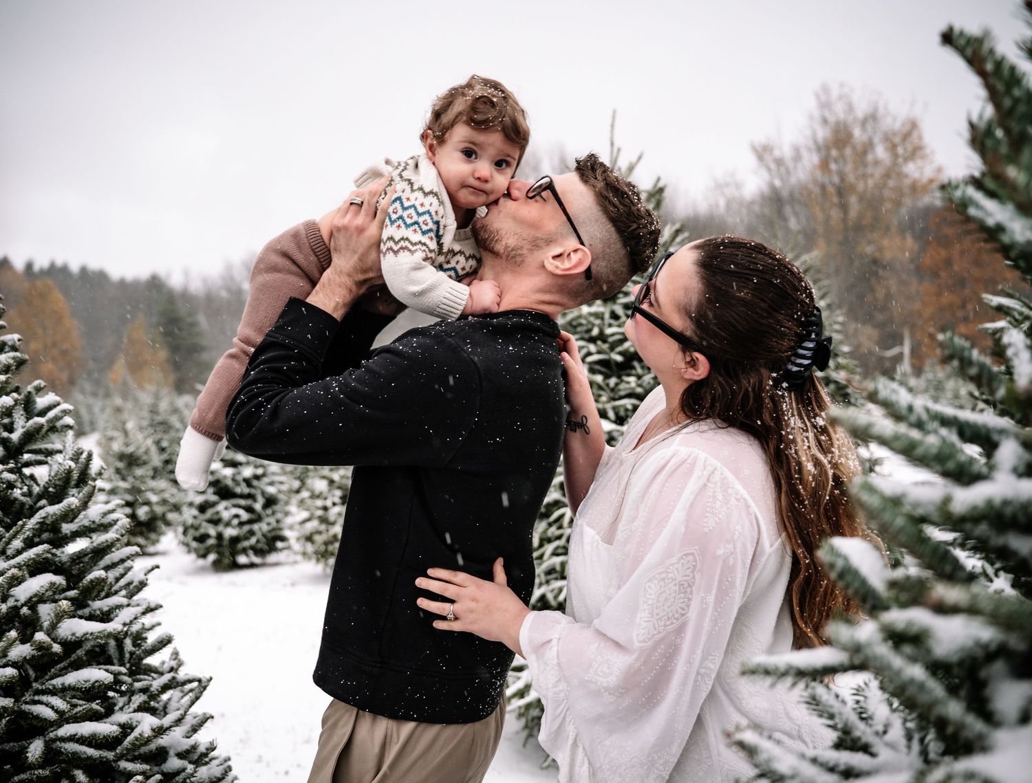 A couple shares a romantic moment in a snowy winter landscape with evergreen trees in the background.