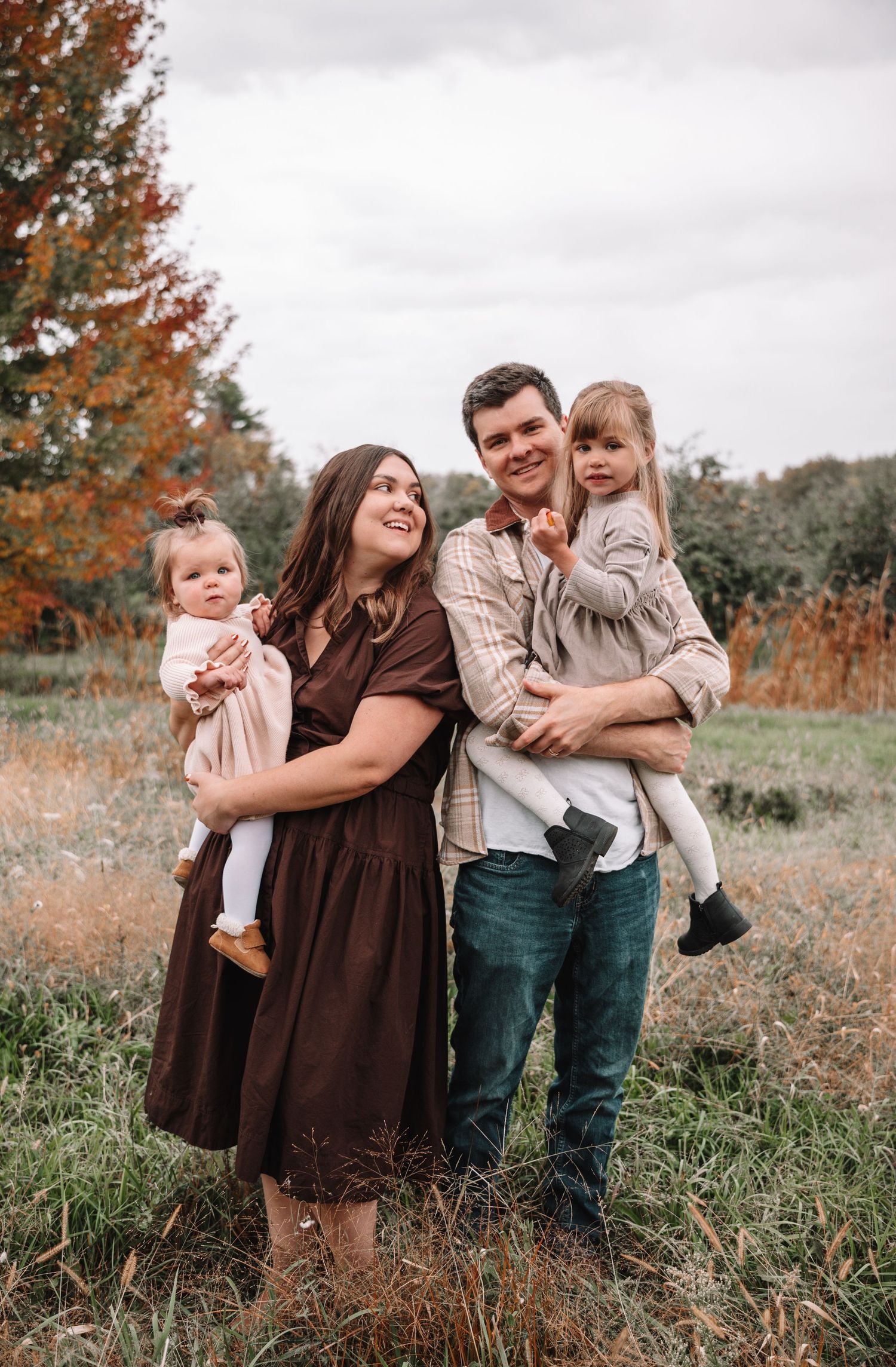 A family laughs and plays together in a field during autumn with orange foliage in the background.