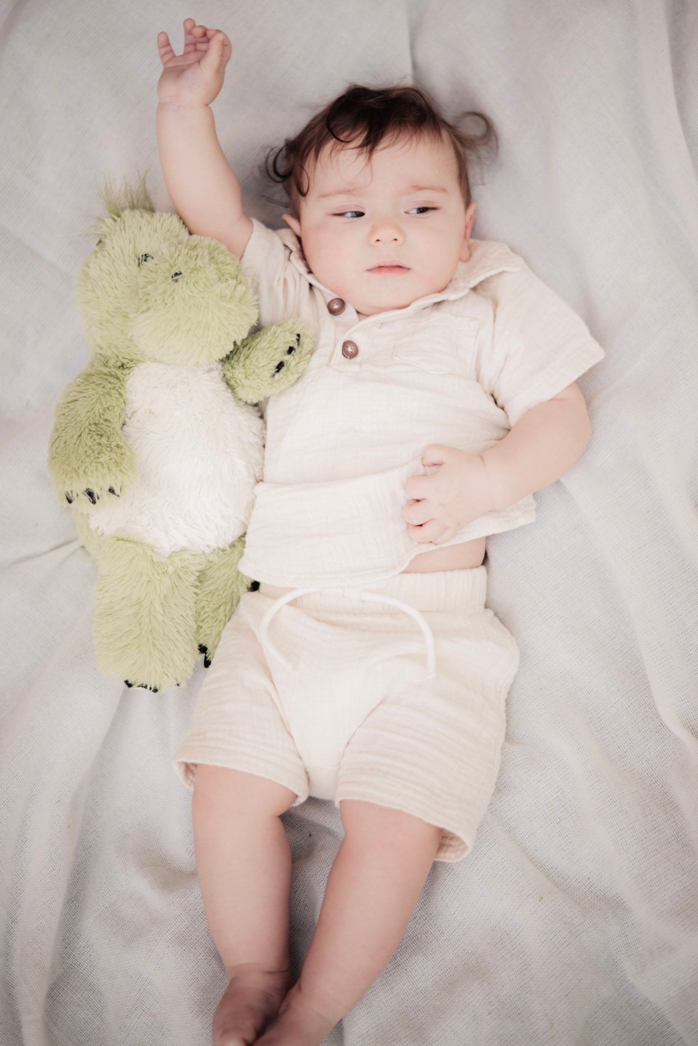 Baby in light colored outfit lying on white fabric with plush green toy.