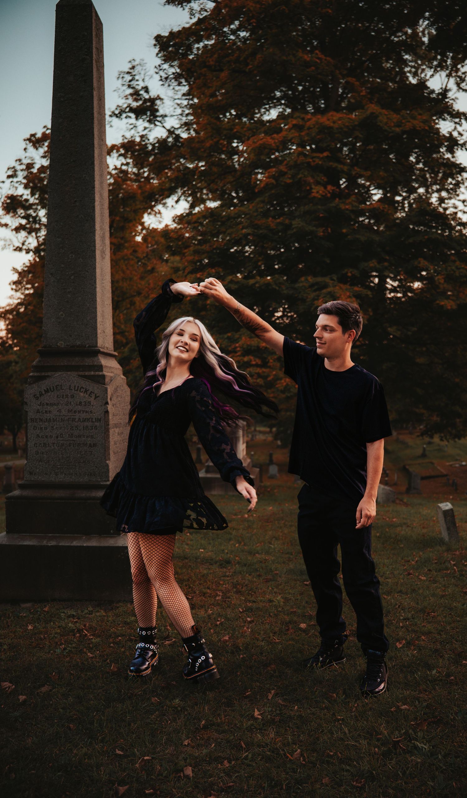 A couple in dark clothing dancing playfully among gravestones at sunset in an autumn cemetery.