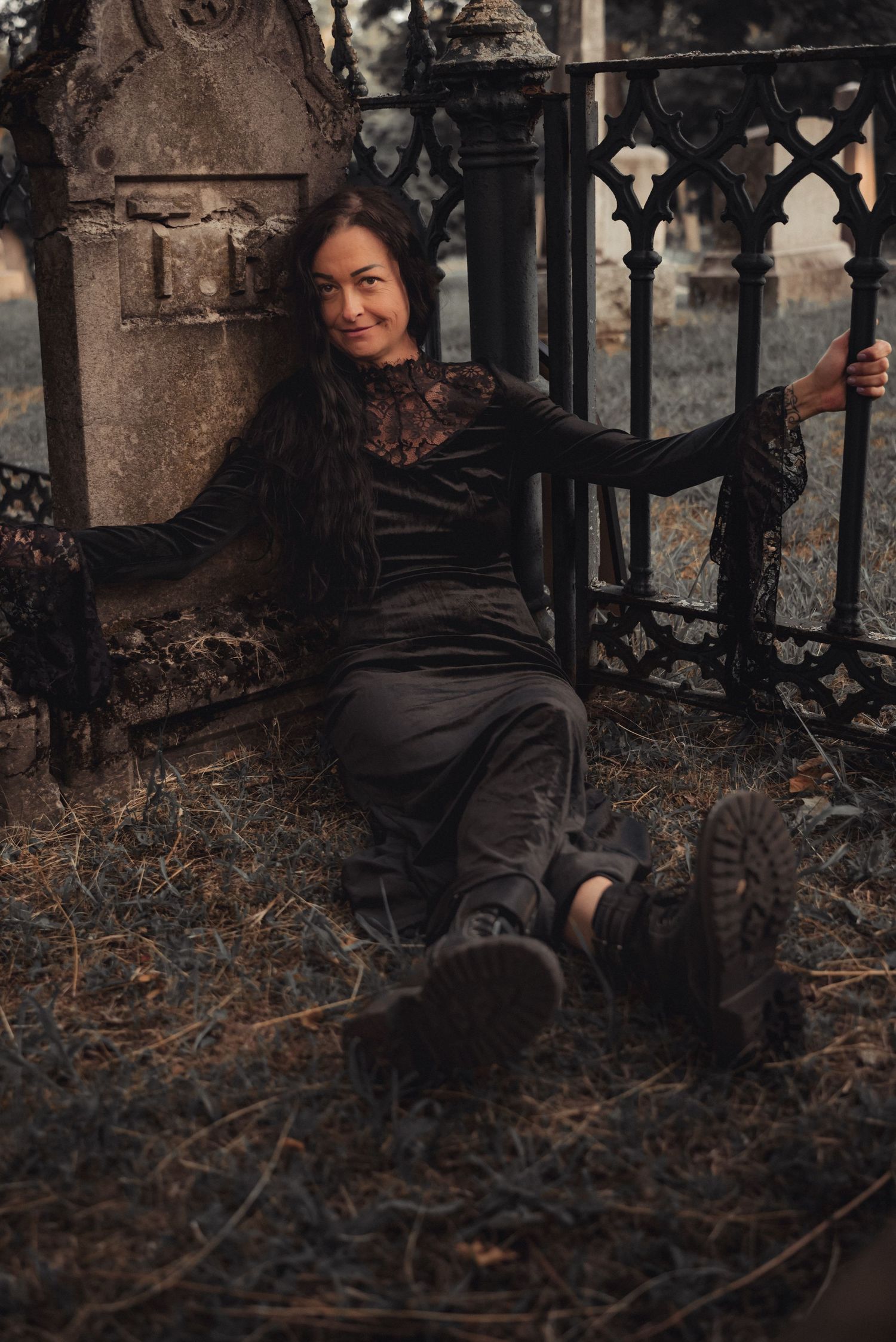 A figure in a black gothic dress sits on stone steps beside ornate iron railings in a moody autumn setting.