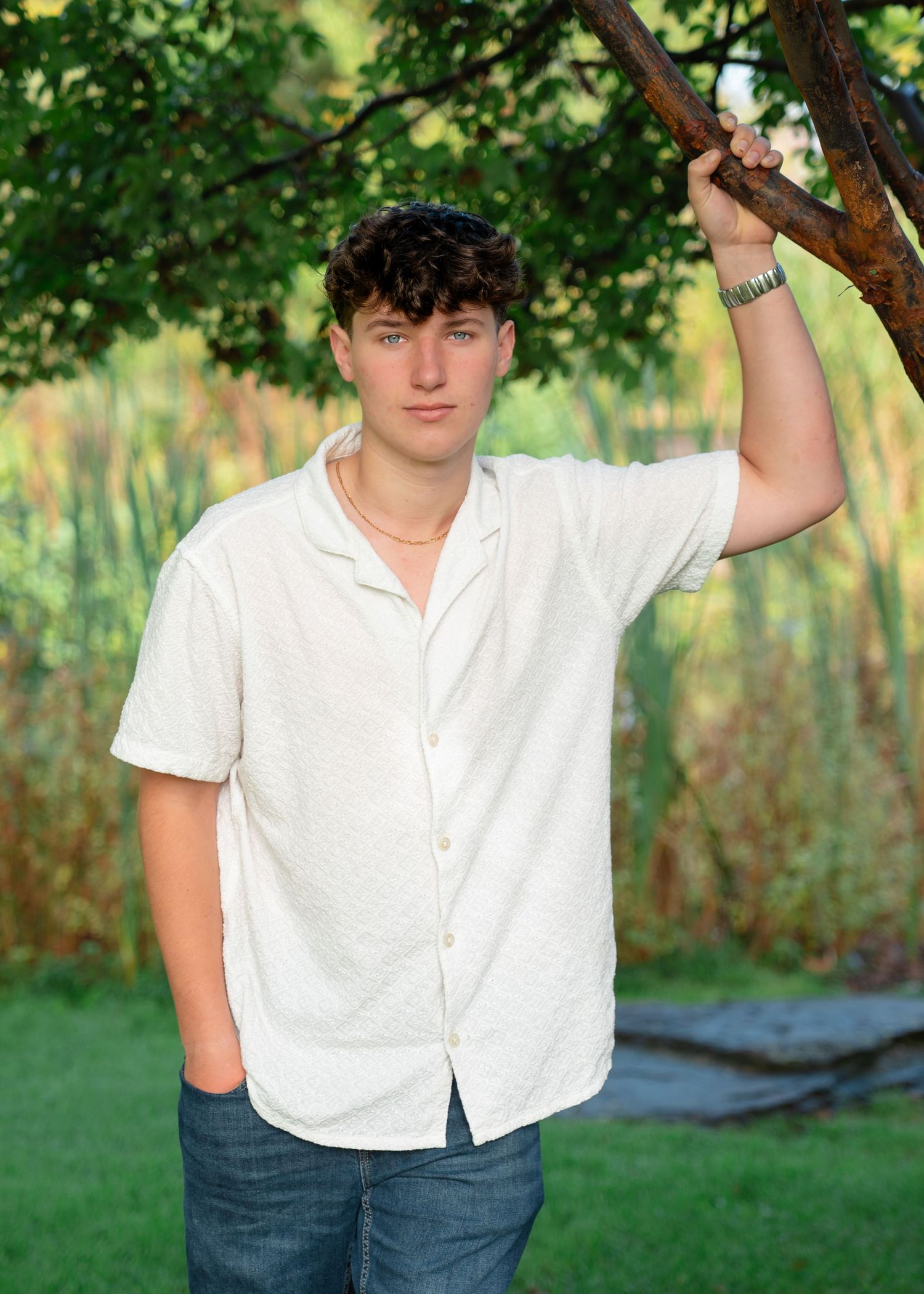 A person in a white button-down shirt and jeans poses casually against a tree in a lush green outdoor setting.
