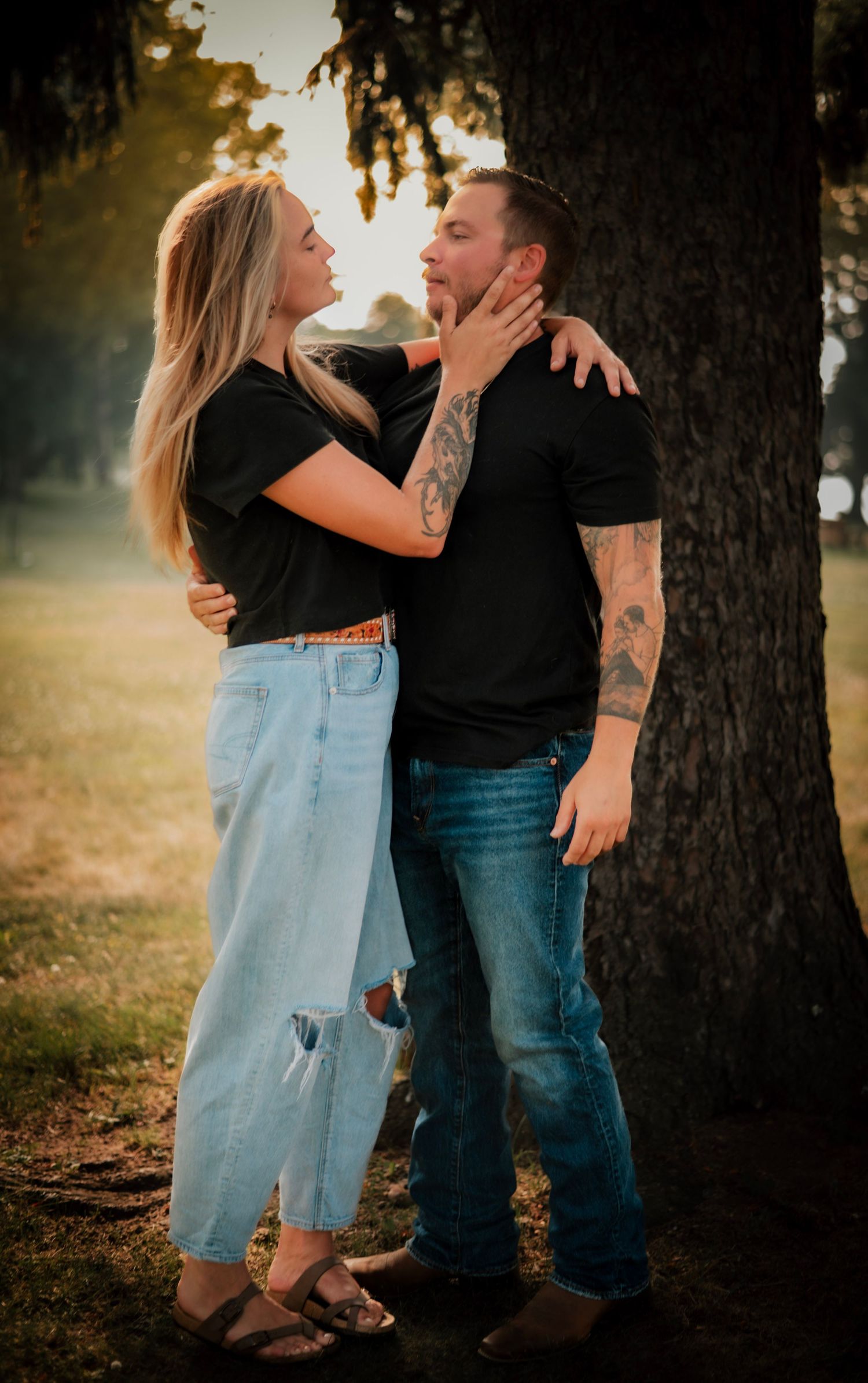 Romantic couple in casual attire sharing intimate moments against a sunlit tree backdrop in a park setting.