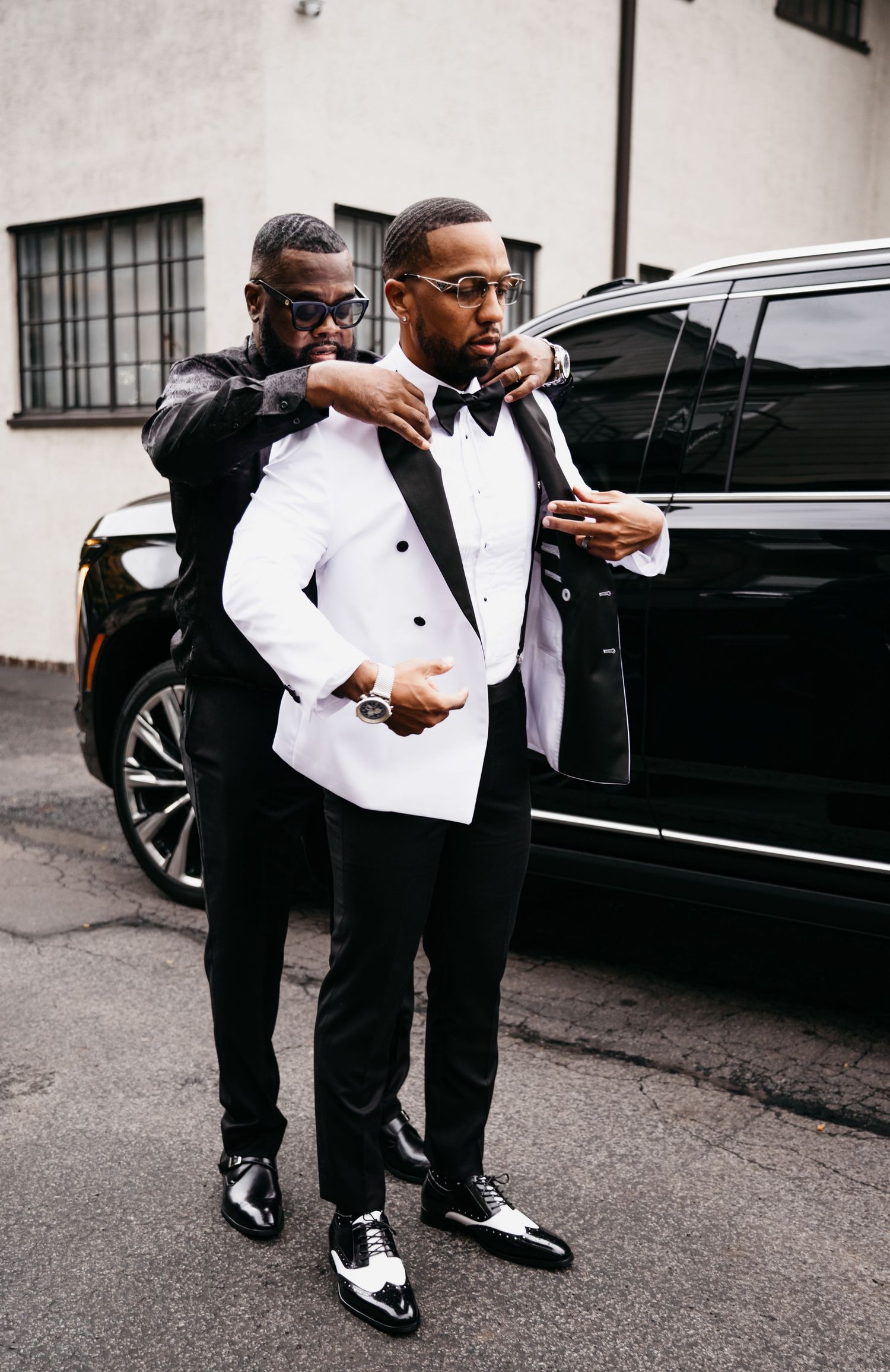 A group in formal black and white attire pose playfully near a luxury black vehicle on a city street.