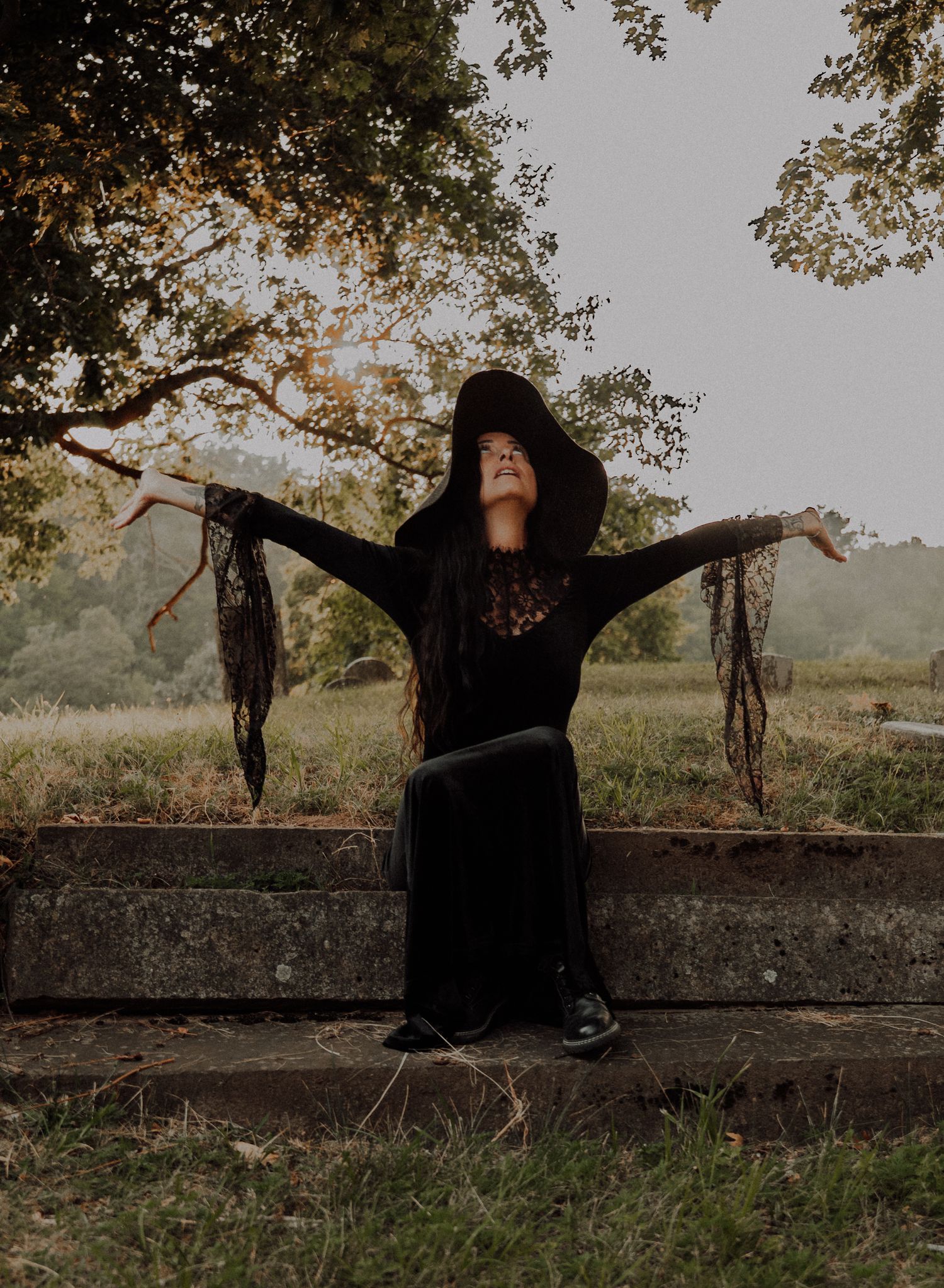 A figure in black clothing sits and poses expressively against a stone wall in a natural outdoor setting at sunset.