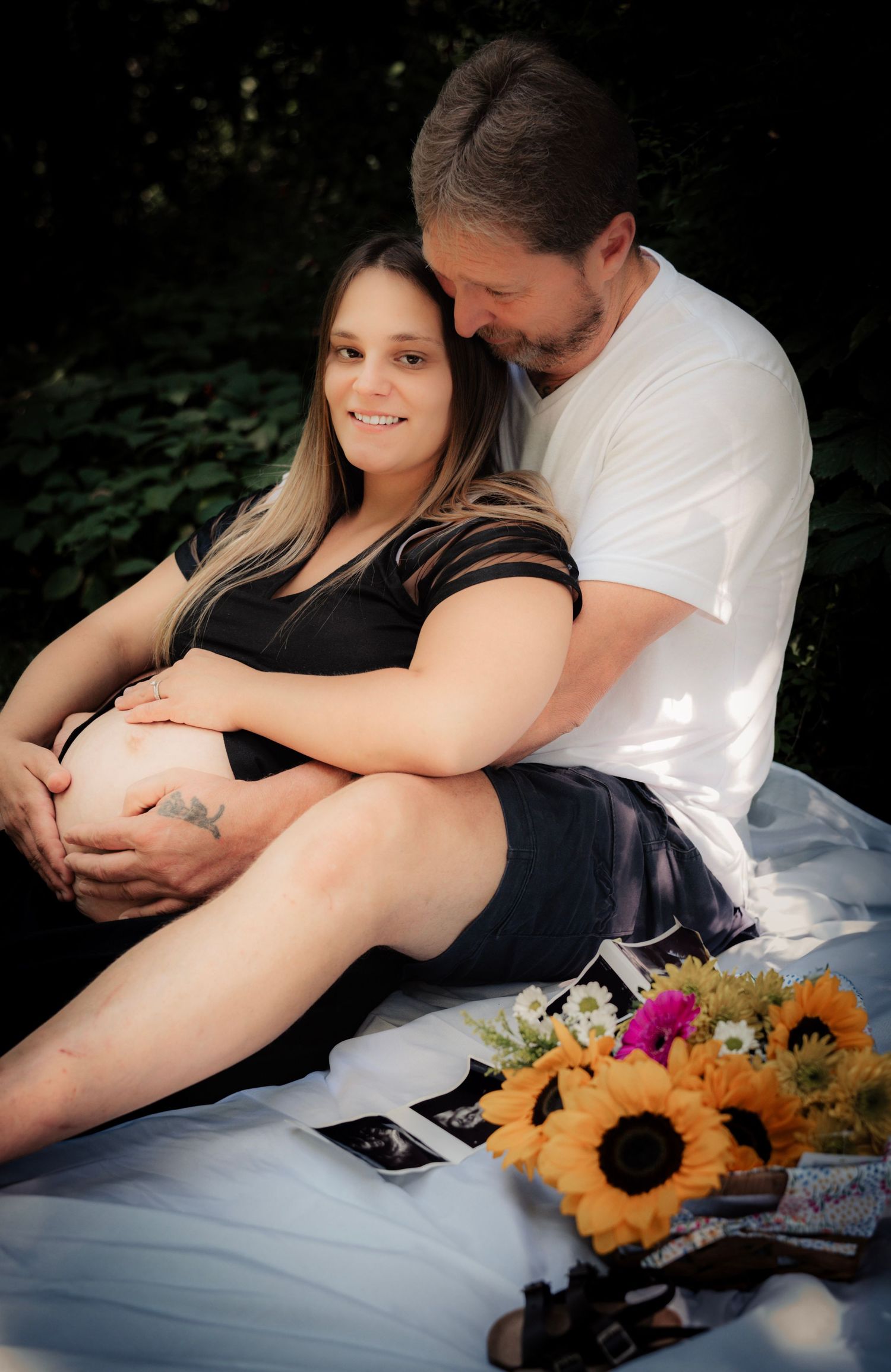 A romantic maternity photo session with sunflowers showing a couple embracing on a white blanket outdoors.