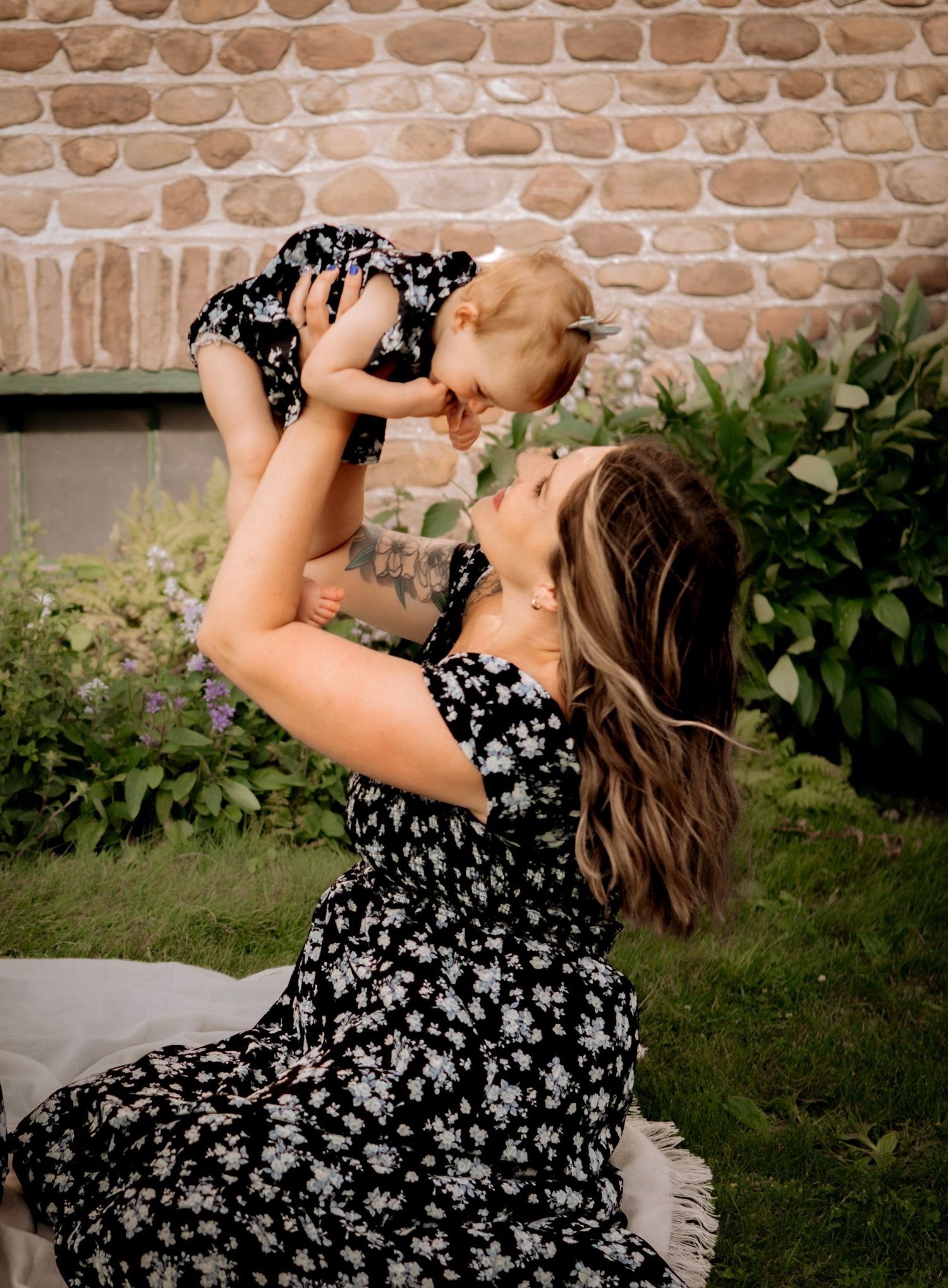 Woman in floral dress lifts baby joyfully in garden setting.