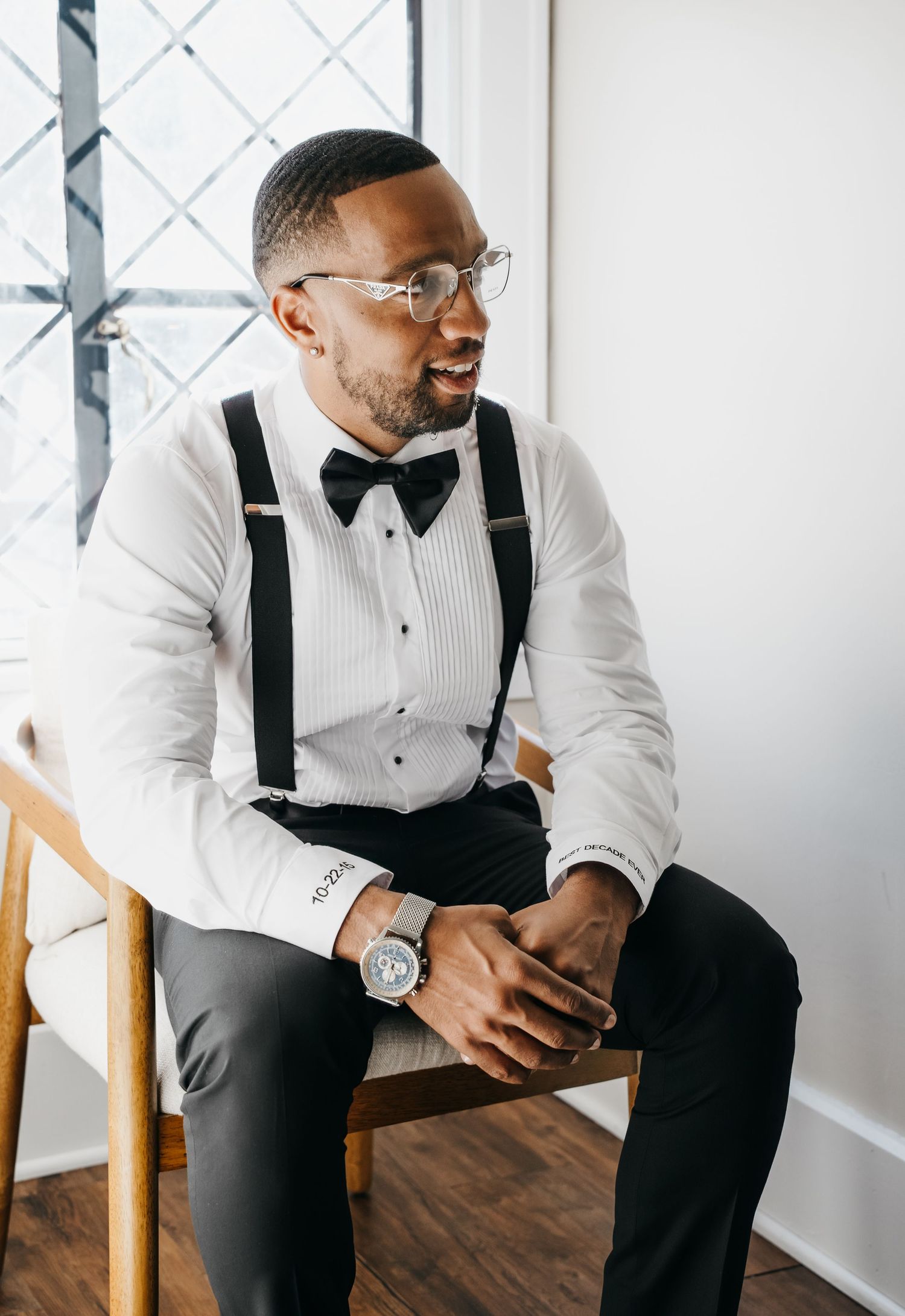 Stylish groom in white tuxedo shirt and black suspenders sitting on wooden chair in bright room.