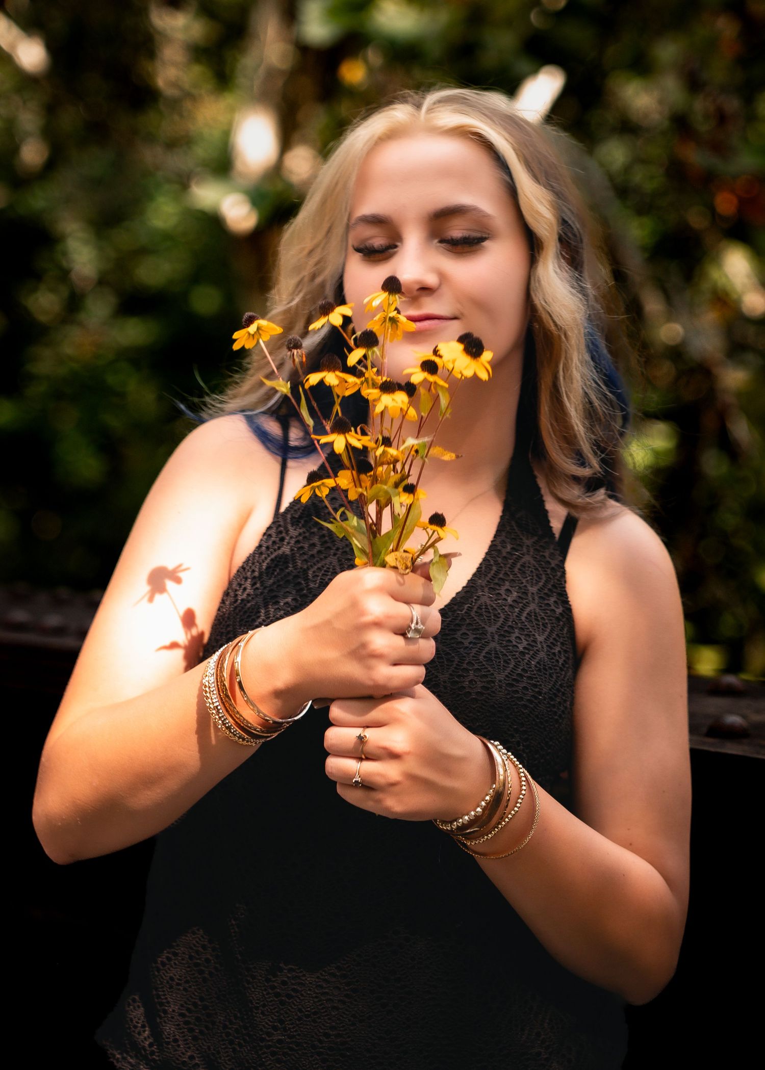Someone in a black sleeveless top smells yellow wildflowers while wearing gold bangles in a sunlit outdoor setting.