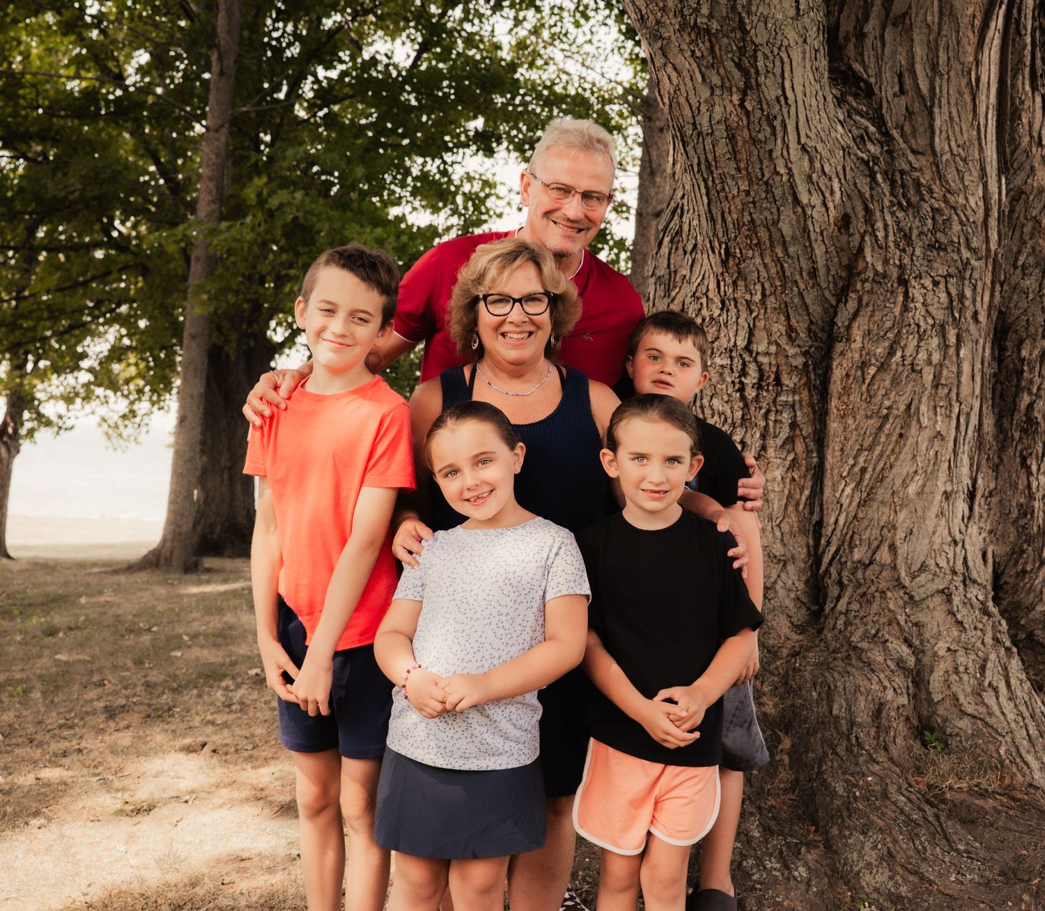 A group gathers for a casual family photo under large trees in a park setting.