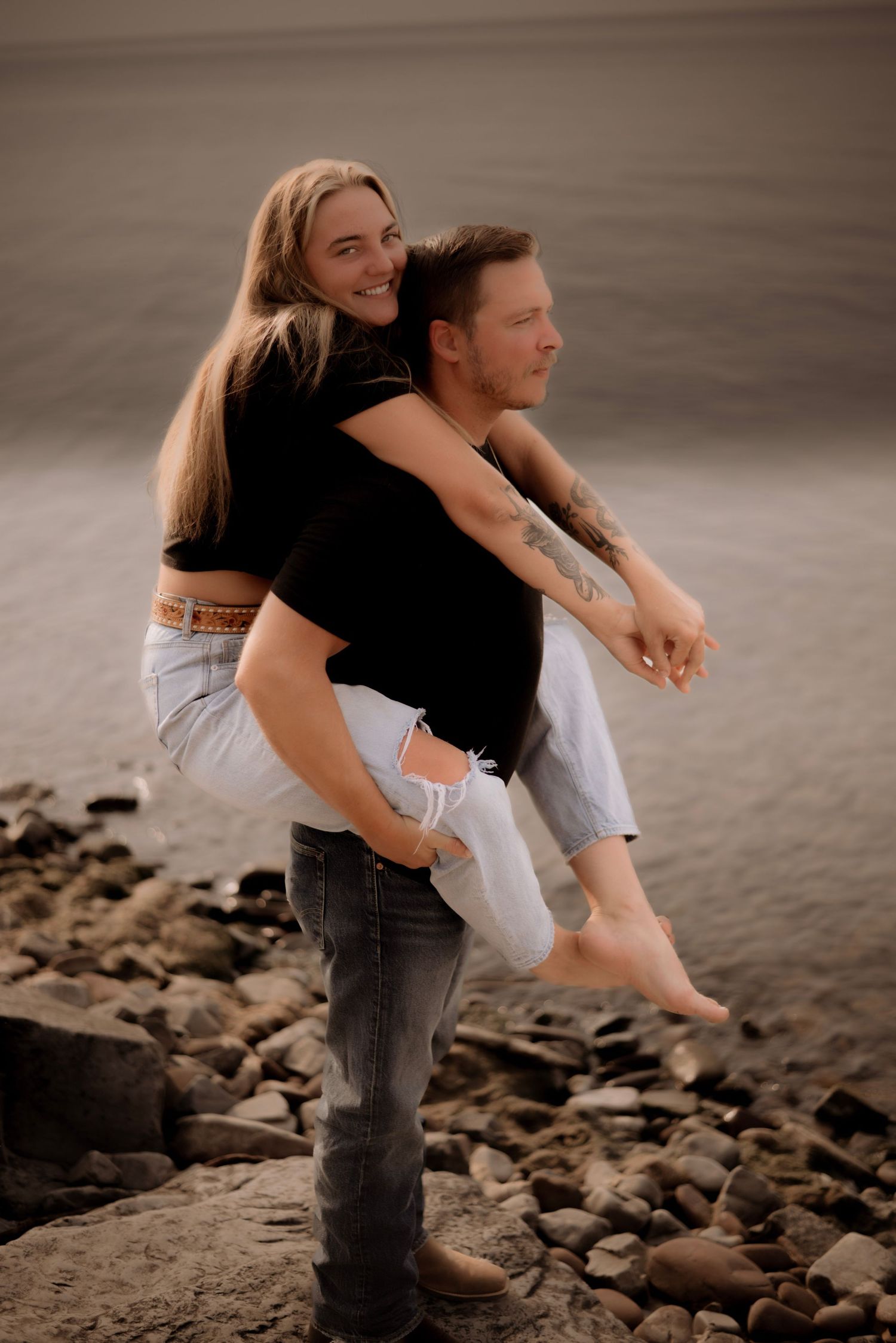 Couple embracing on rocky shoreline during golden hour creating intimate silhouettes against muted background.