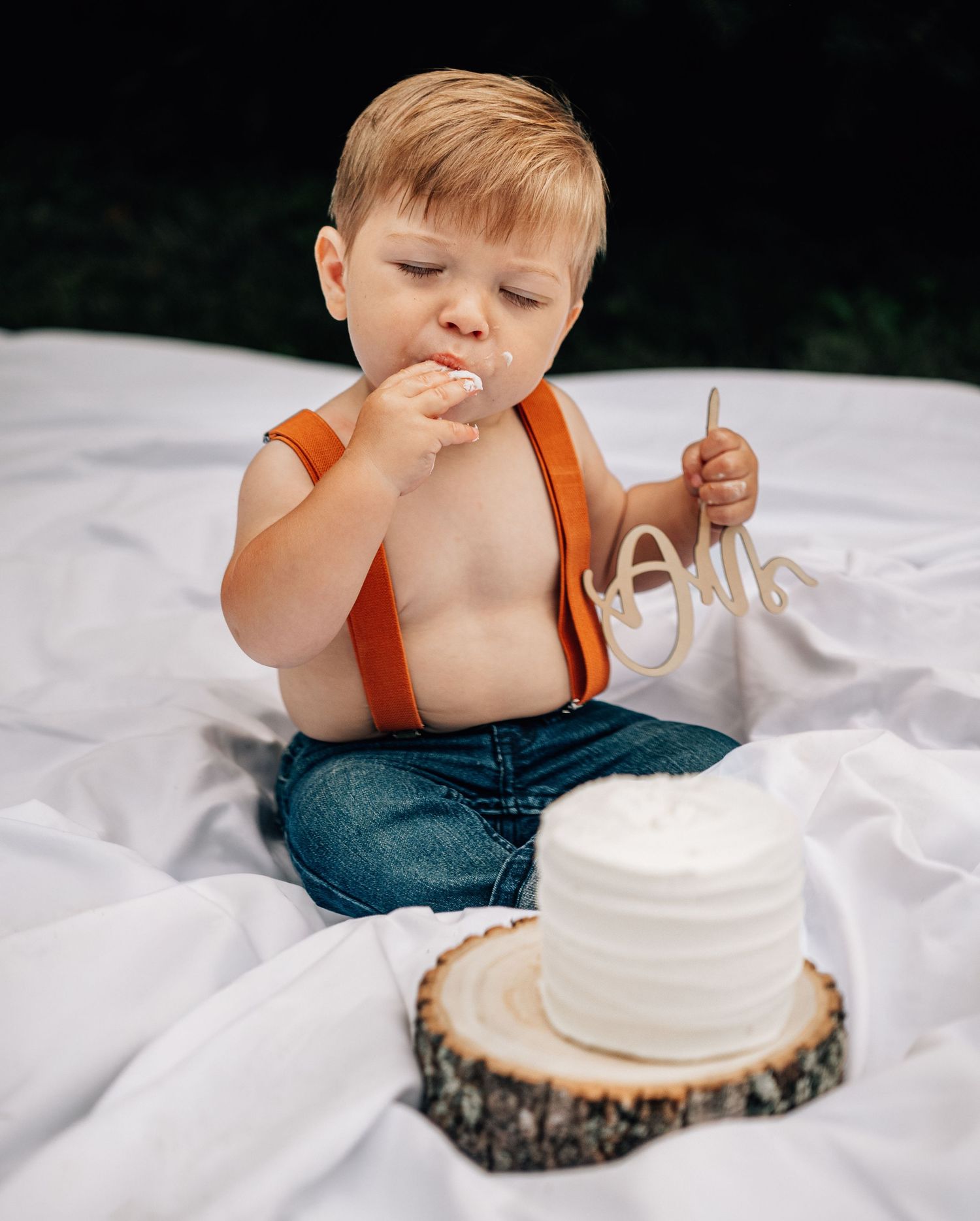 A small child in jeans and orange top sitting on white bedding while reaching towards a white frosted cake.