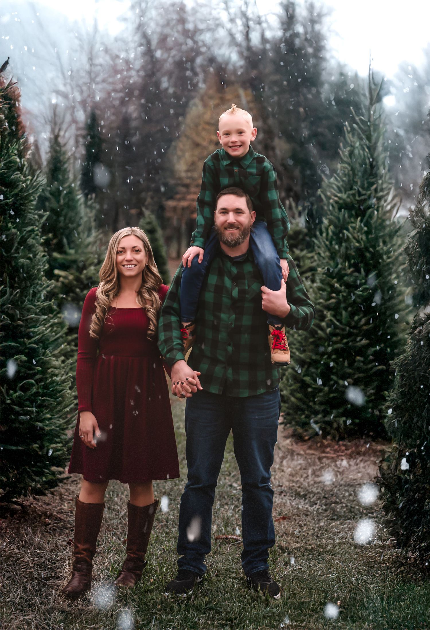 A couple poses together at a Christmas tree farm in winter while snow falls around them wearing plaid and burgundy outfits.
