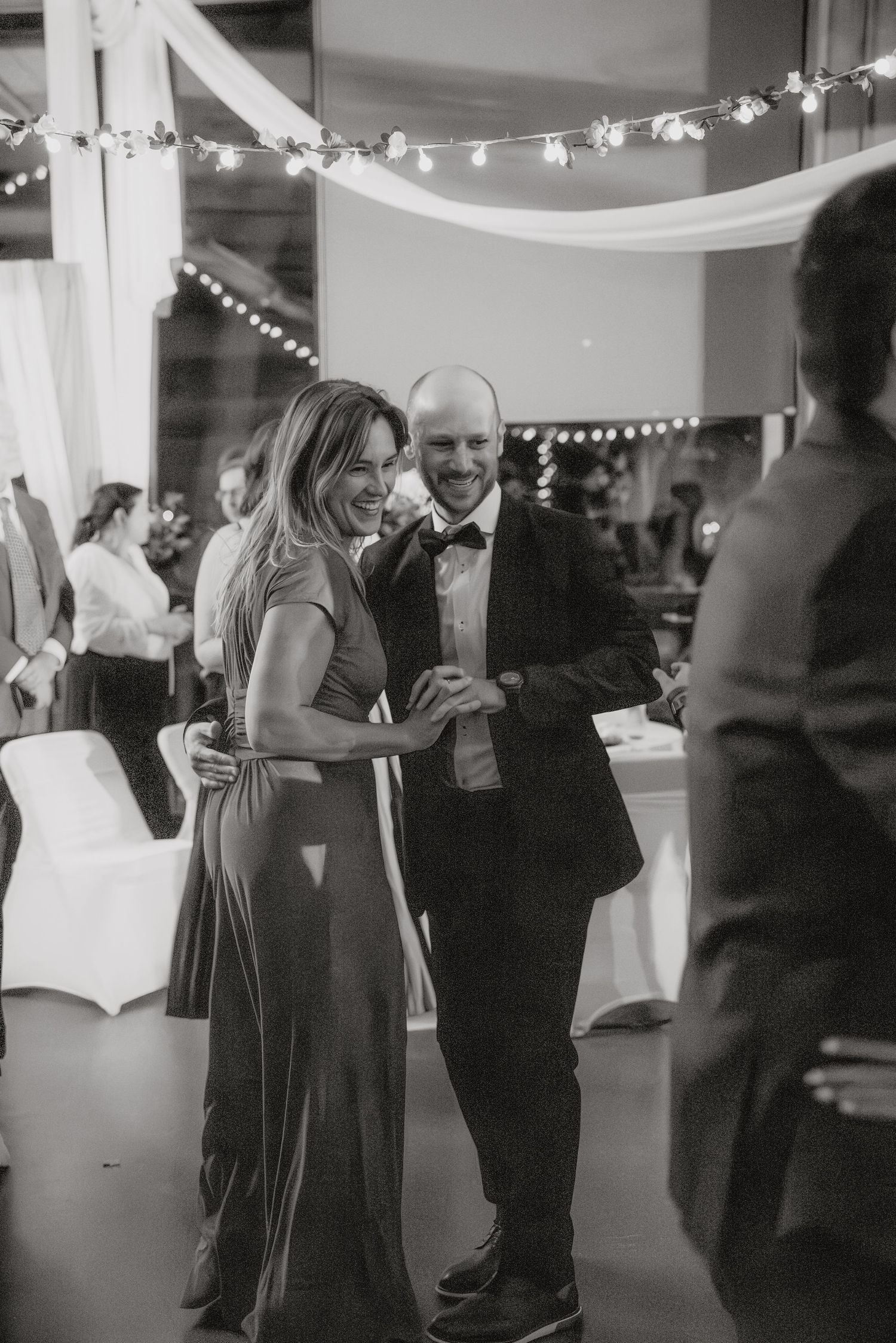Wedding guests dance together at an indoor reception decorated with string lights and white fabric draping.