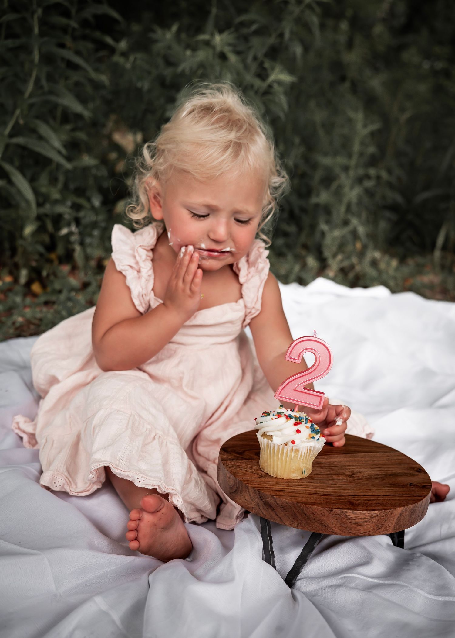 Toddler in pink dress sits on white blanket with birthday cupcake and number 2 candle on wooden table.