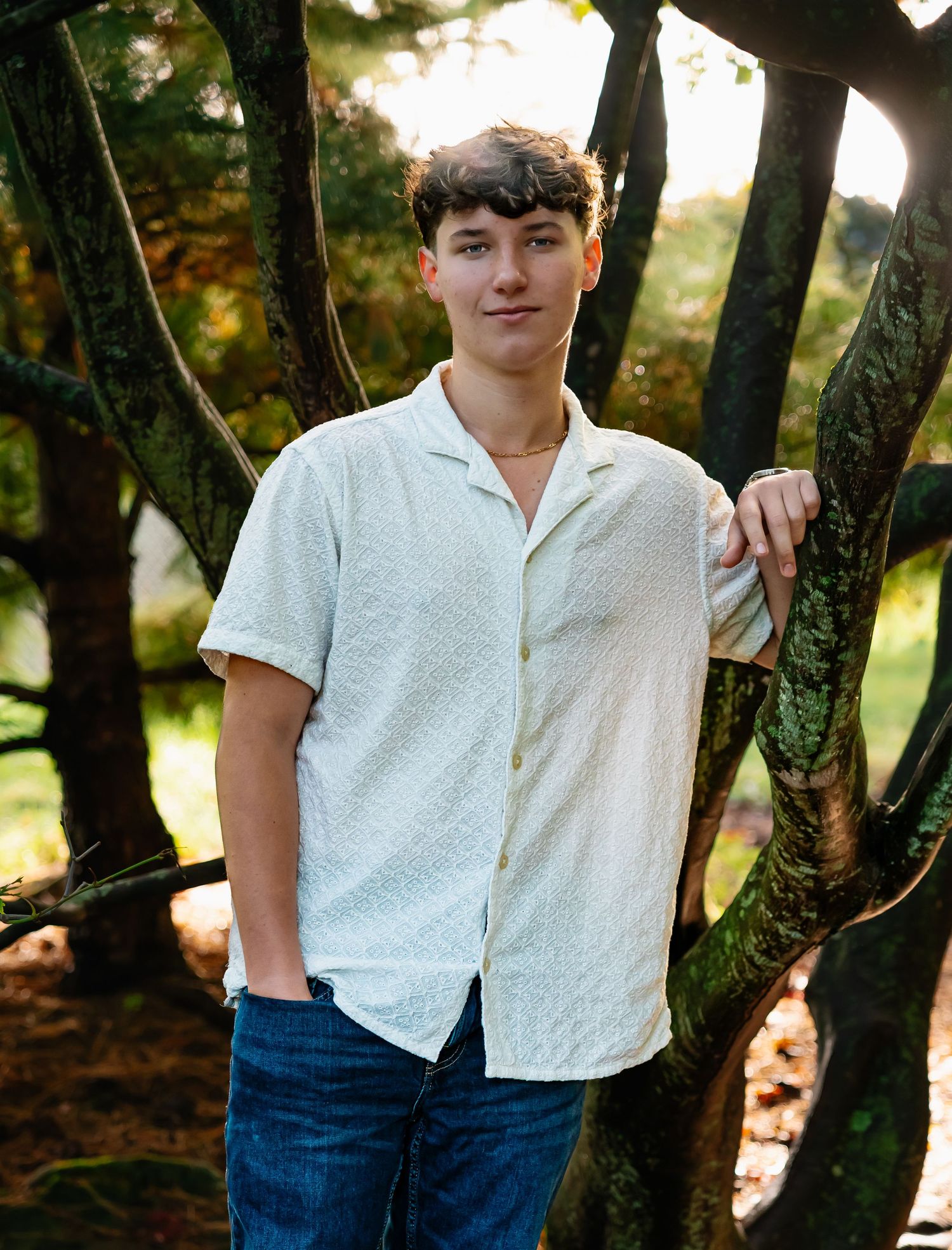 A person wearing a white button-down shirt and jeans poses casually against tree trunks in a natural outdoor setting.