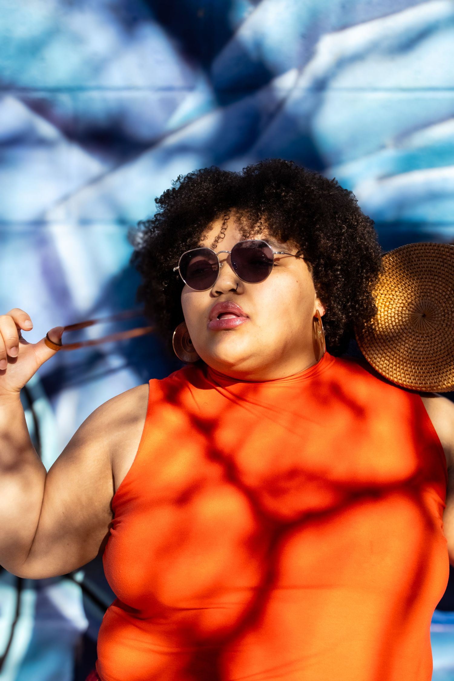 Figure in orange top and sunglasses against blue background with dramatic lighting.