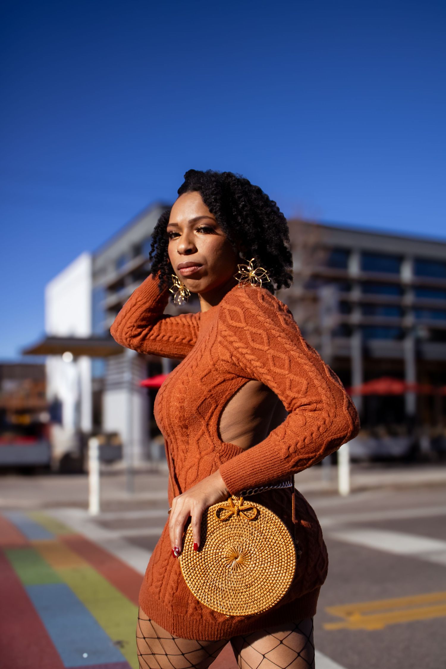 Fashion model poses in bright orange sweater and woven straw handbag against urban backdrop with rainbow crosswalk.